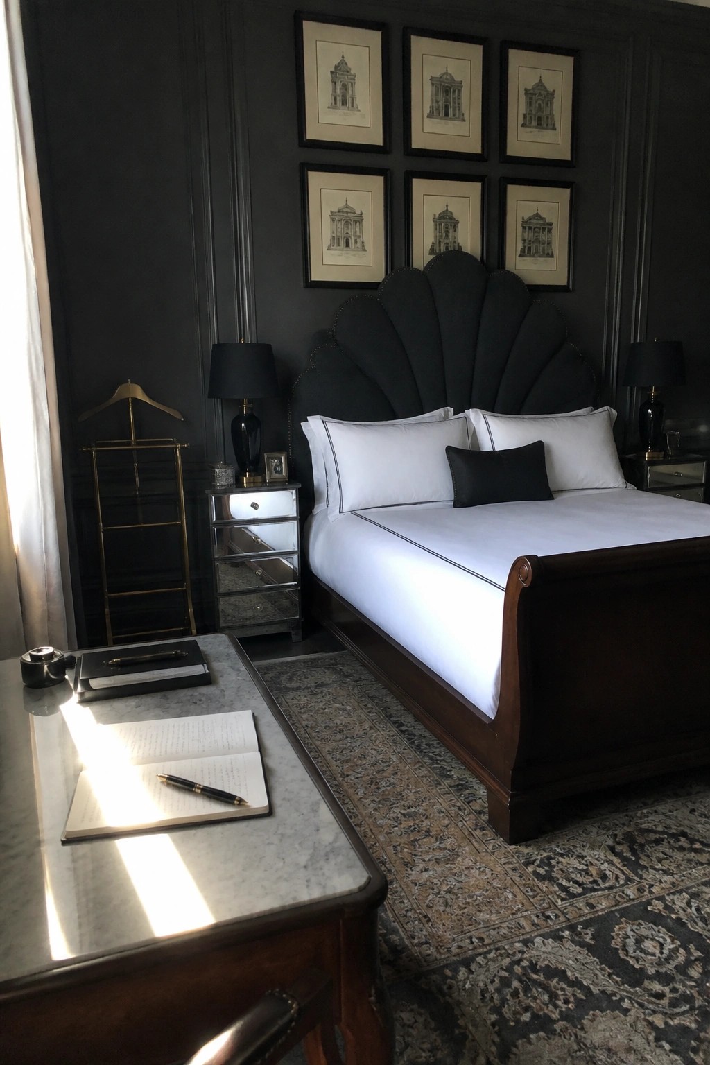 A dark bedroom with a large black upholstered shell-shaped headboard on a wooden sleigh bed, white linens, framed art above, and a marble desk in the foreground.
