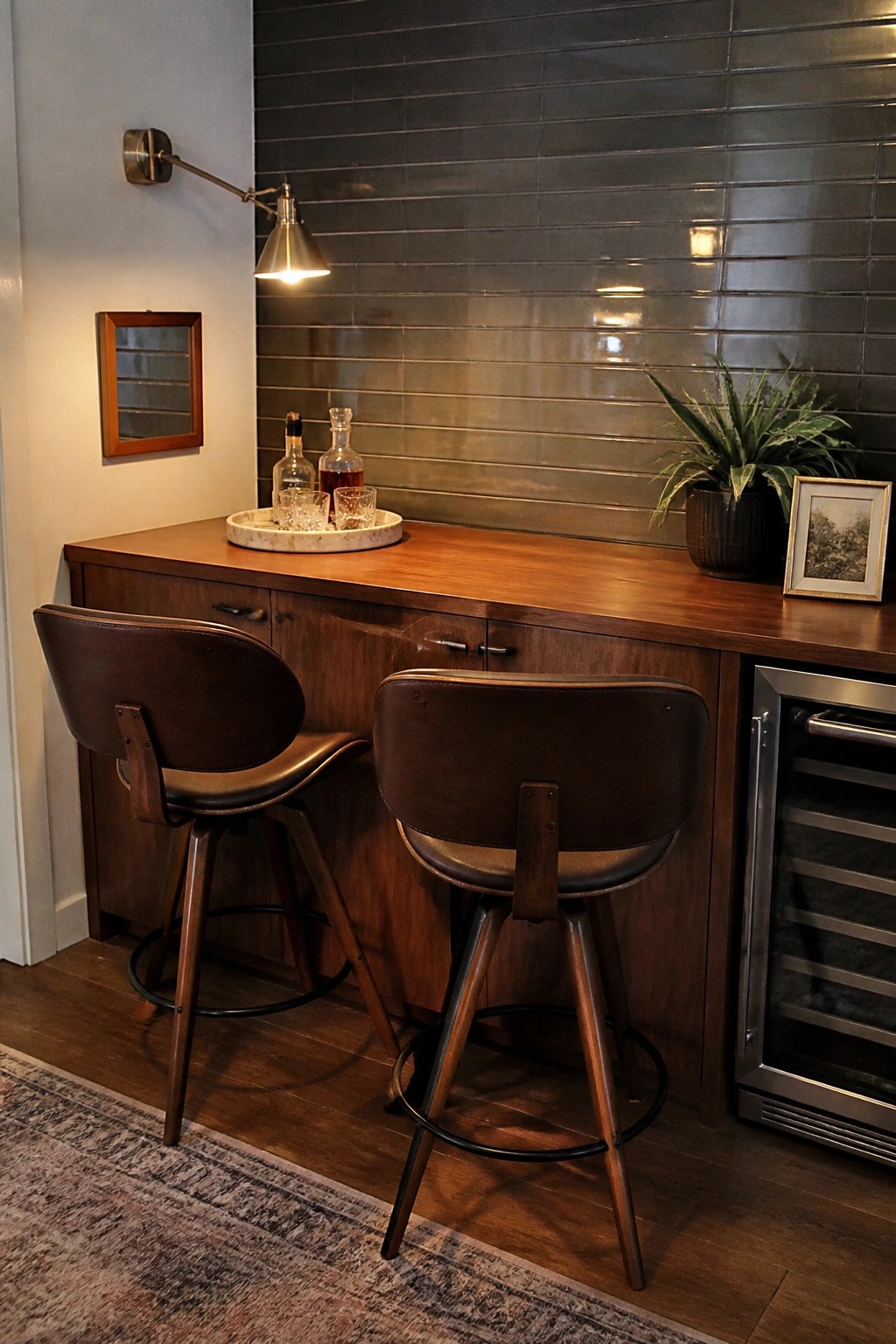 Two dark leather bar stools face a long wooden counter with cabinet storage below, a marble tray holding glassware, and a wine fridge at the far end.