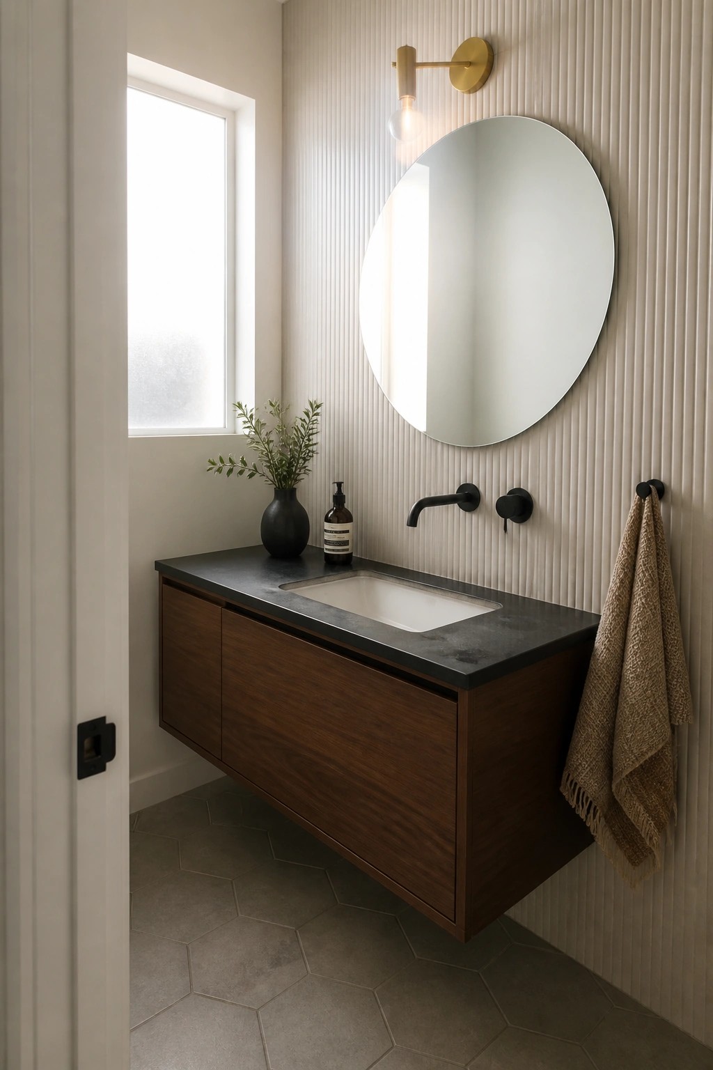 A bathroom with a floating dark wood vanity, dark stone countertop, white undermount sink, round wall mirror, and black wall-mounted faucet and towel hook.