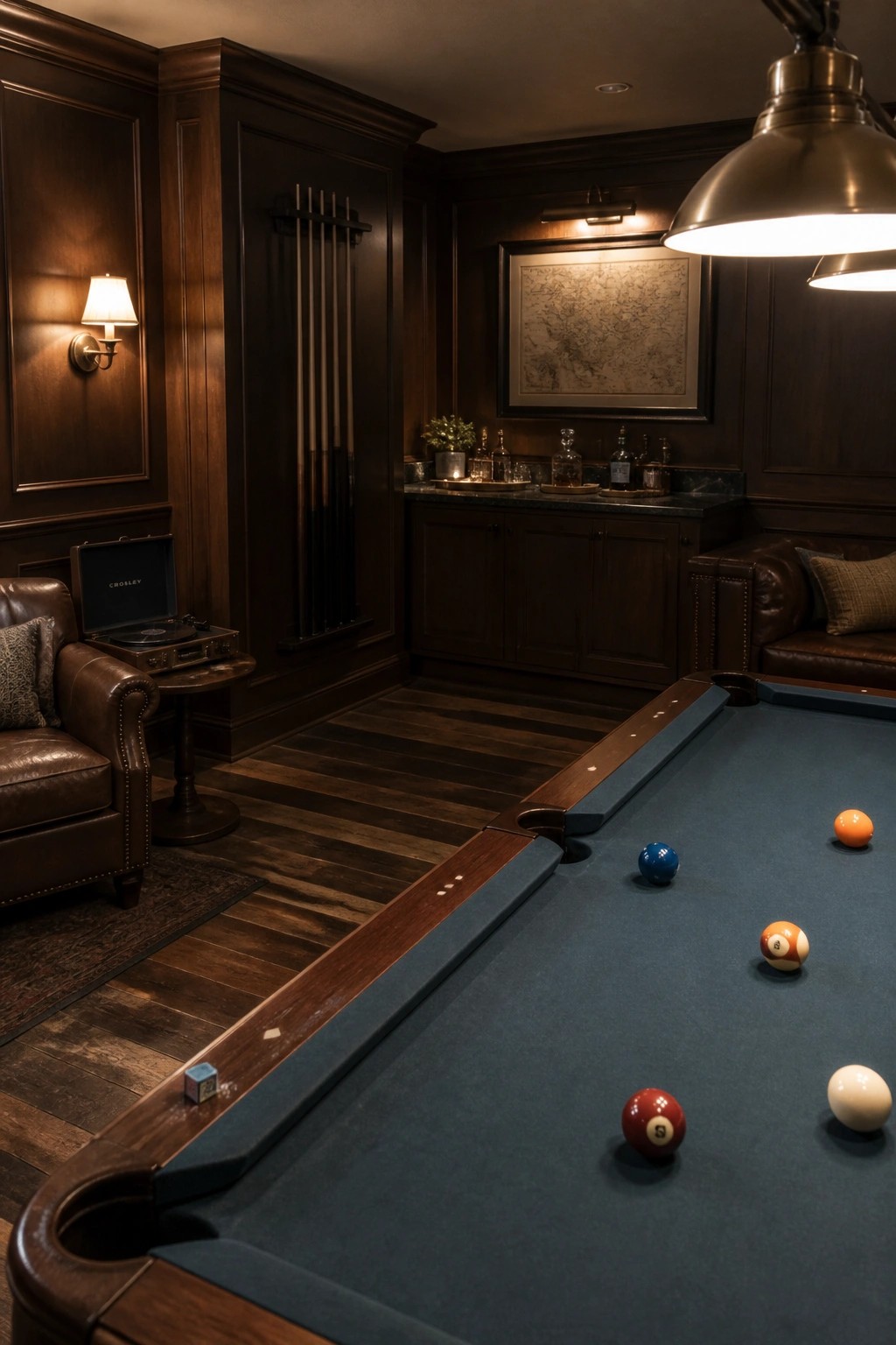 Interior of a masculine billiards room featuring dark wood paneling, a blue felt pool table, leather armchairs, and a built-in bar area with cabinets.