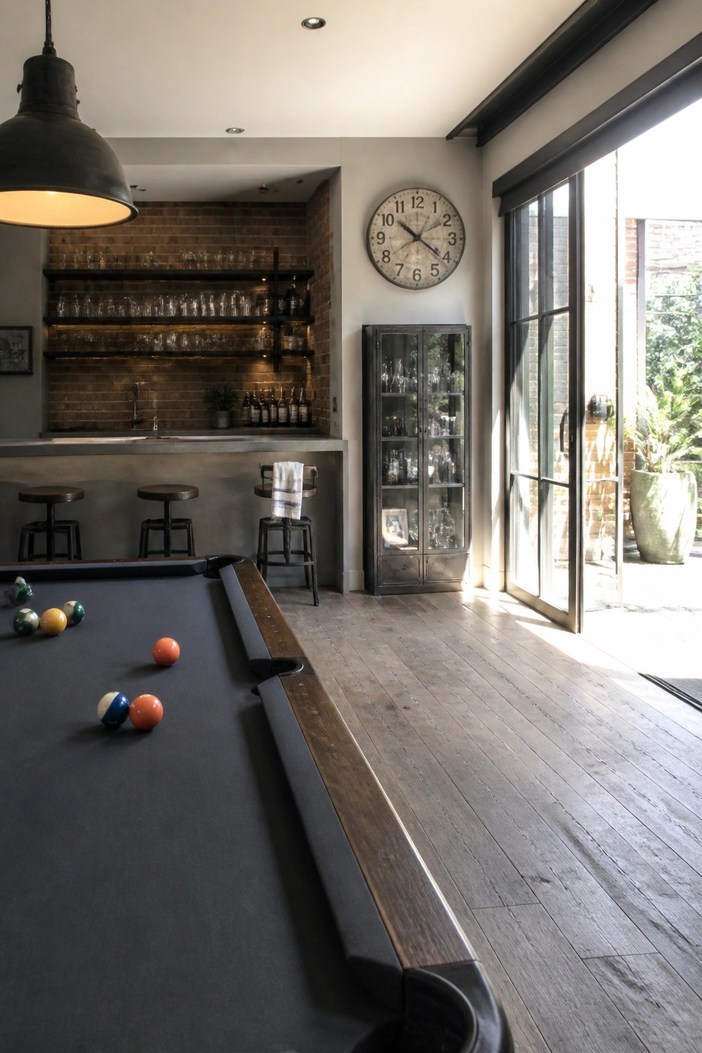 A basement game room with a pool table in the foreground, a concrete bar counter with three stools, open shelving on an exposed brick wall, and a tall metal cabinet.