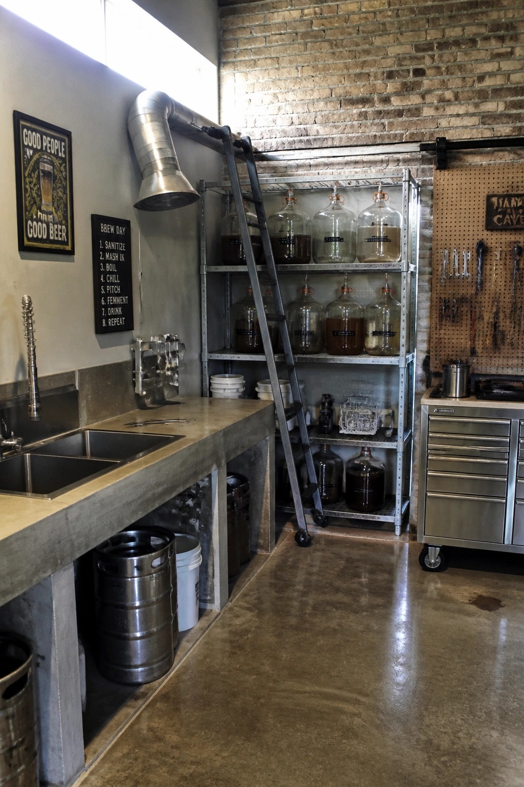 Interior view of a home brewery with concrete countertops, double stainless sinks, tall rolling metal shelves holding glass carboys, and an exposed brick wall.