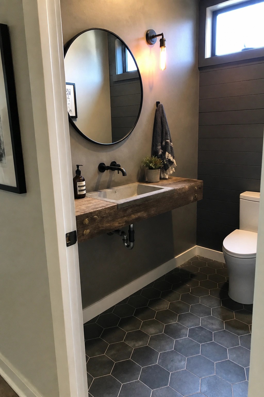 A bathroom interior showing a floating reclaimed wood vanity with a concrete sink, round mirror above it, dark metal faucet and wall sconce, and a white toilet on the right.