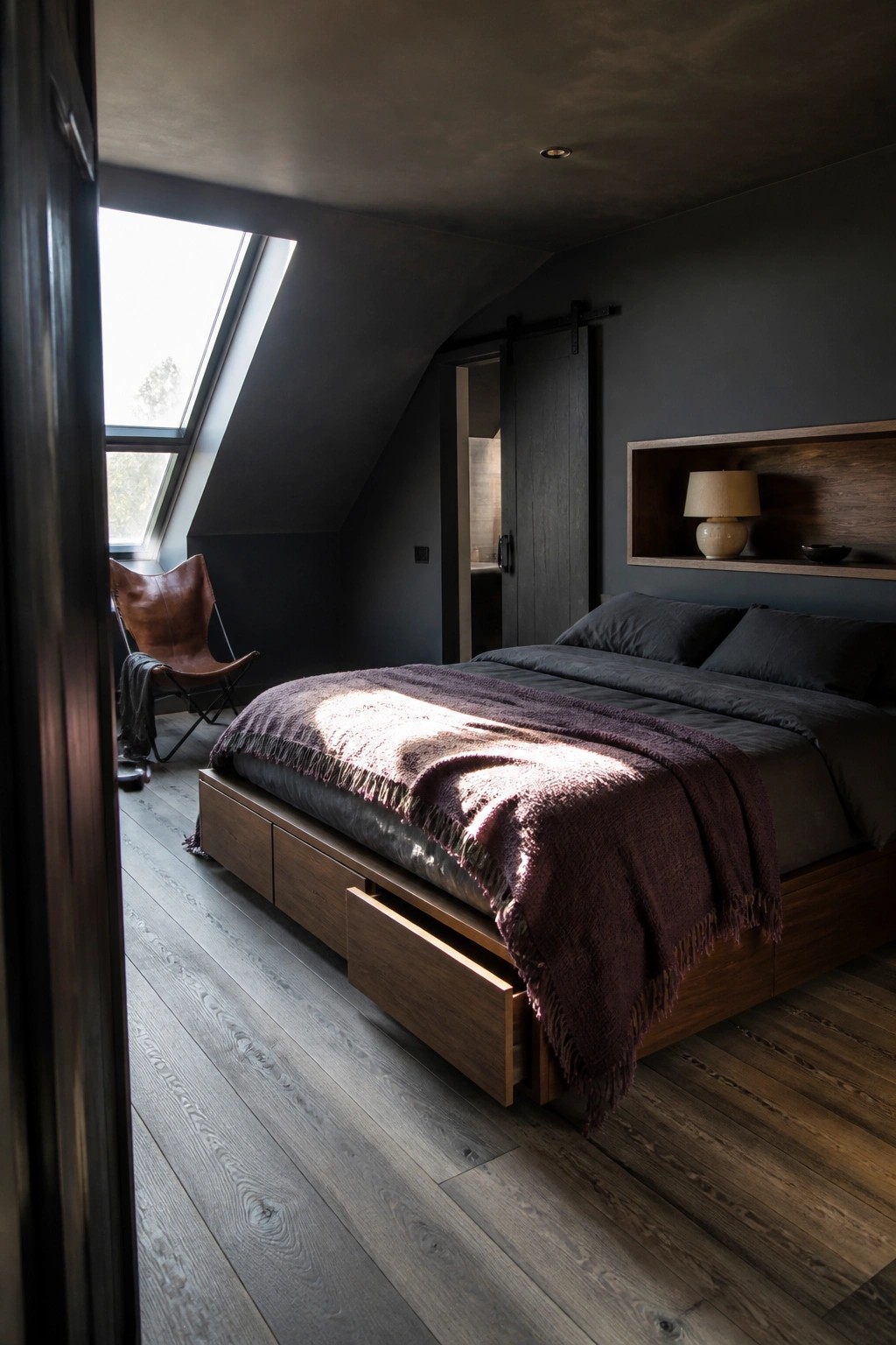 A dark bedroom with a platform bed that has wooden storage drawers built into the base, dark bedding, and a skylight on the slanted ceiling.