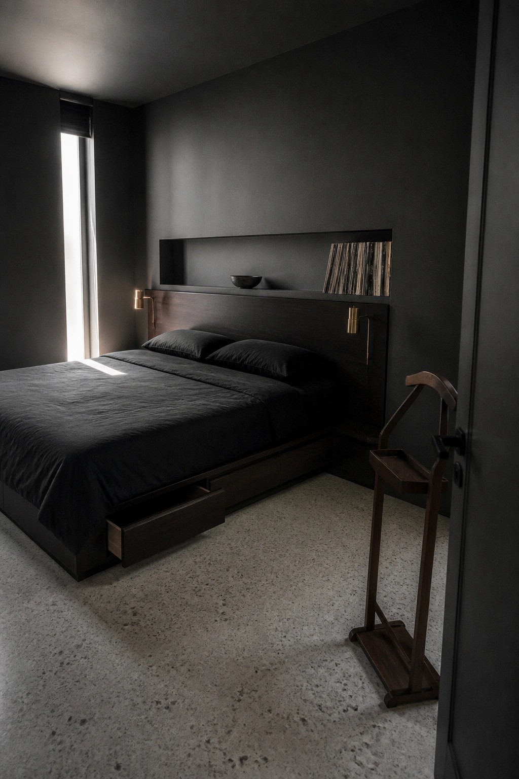 A dark modern bedroom showing a large bed with a wooden headboard, a long recessed shelf above it holding vinyl records and a small bowl, and two wall-mounted brass sconces.