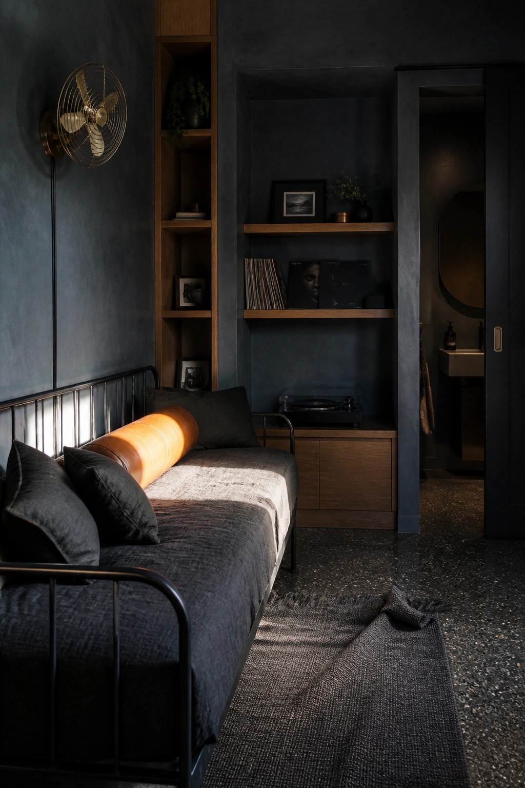 Dark bedroom with daybed, leather bolster, record shelves, brass fan, and open bathroom.