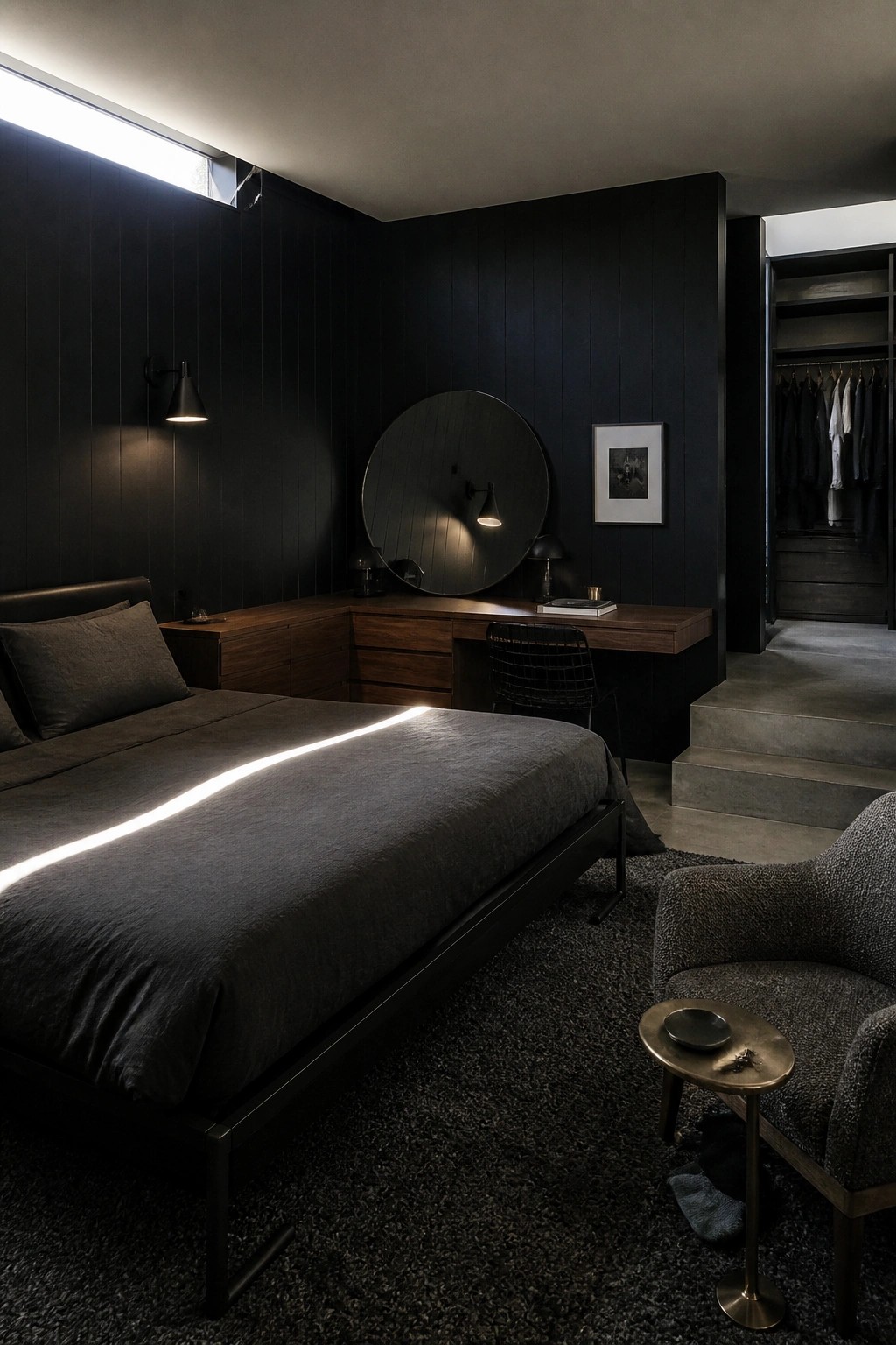 A dark masculine bedroom featuring black vertical wall paneling, a large bed with dark gray bedding, a wooden built-in desk, and a round black mirror.