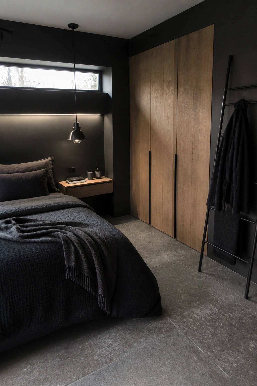 A dark bedroom with black walls, a tall wooden wardrobe with black handles, a bed dressed in dark gray linens, and a black pendant light hanging over a small wooden nightstand.