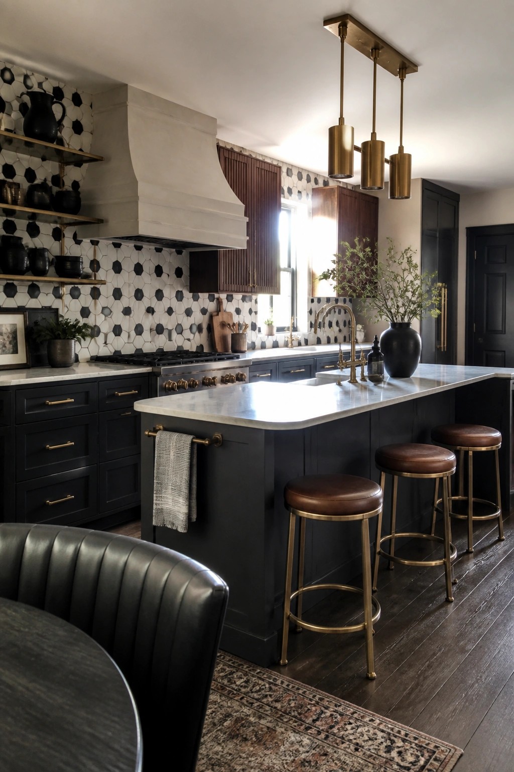 A kitchen interior with a large central island topped in marble, dark navy cabinets, brass pendant lights above three brown leather stools, and a black and white geometric tile backsplash behind the range.