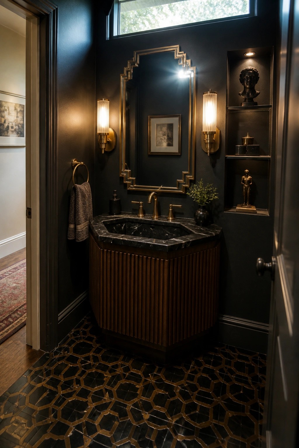 A dark-walled powder room with a fluted wood vanity topped in black marble, brass fixtures, an ornate gold mirror, two ribbed sconces, built-in shelves holding bronze busts, and black and gold geometric floor tiles.