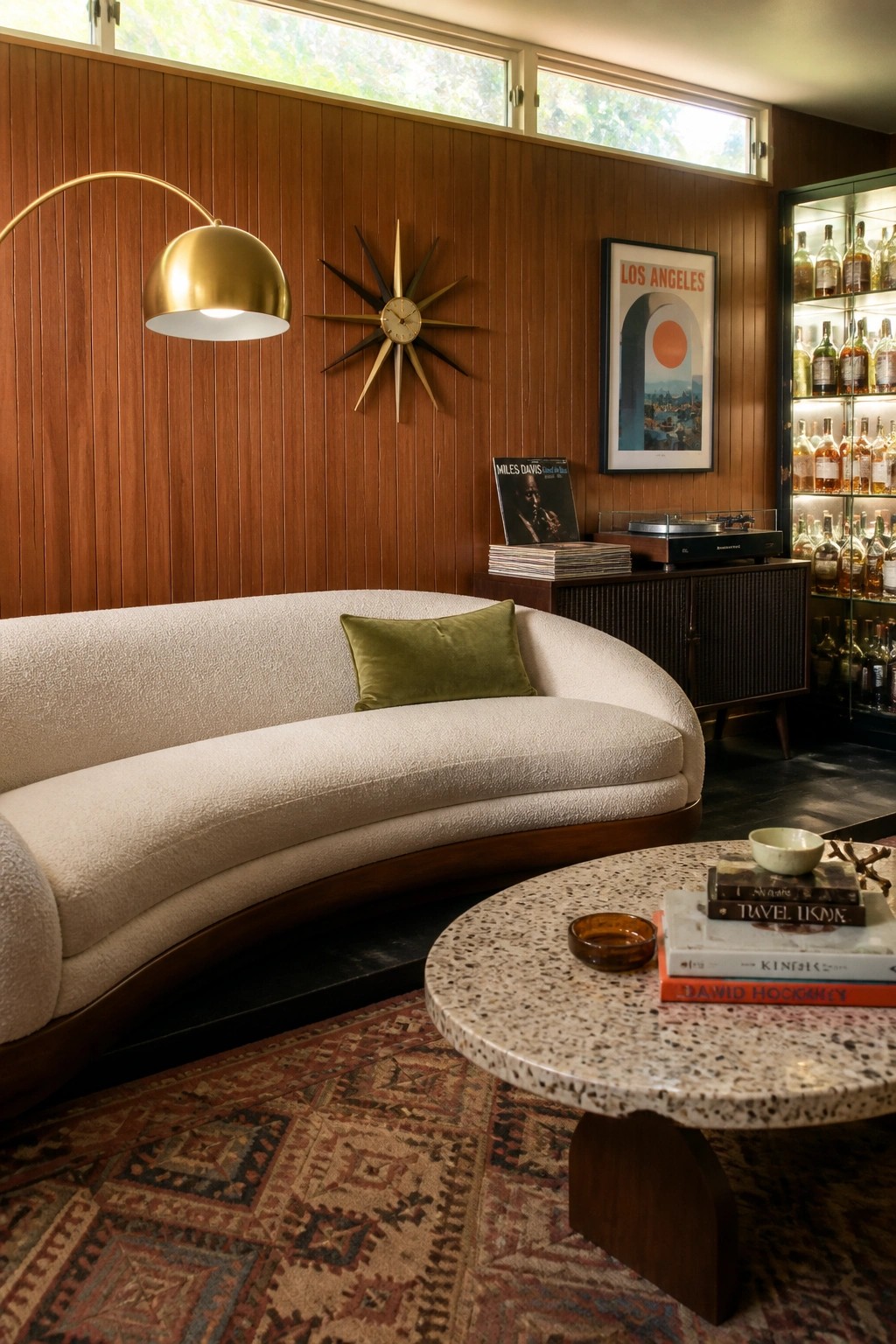 A large curved white sofa with a green pillow faces a terrazzo coffee table in a room with wood paneling, a starburst clock, and a record player setup.