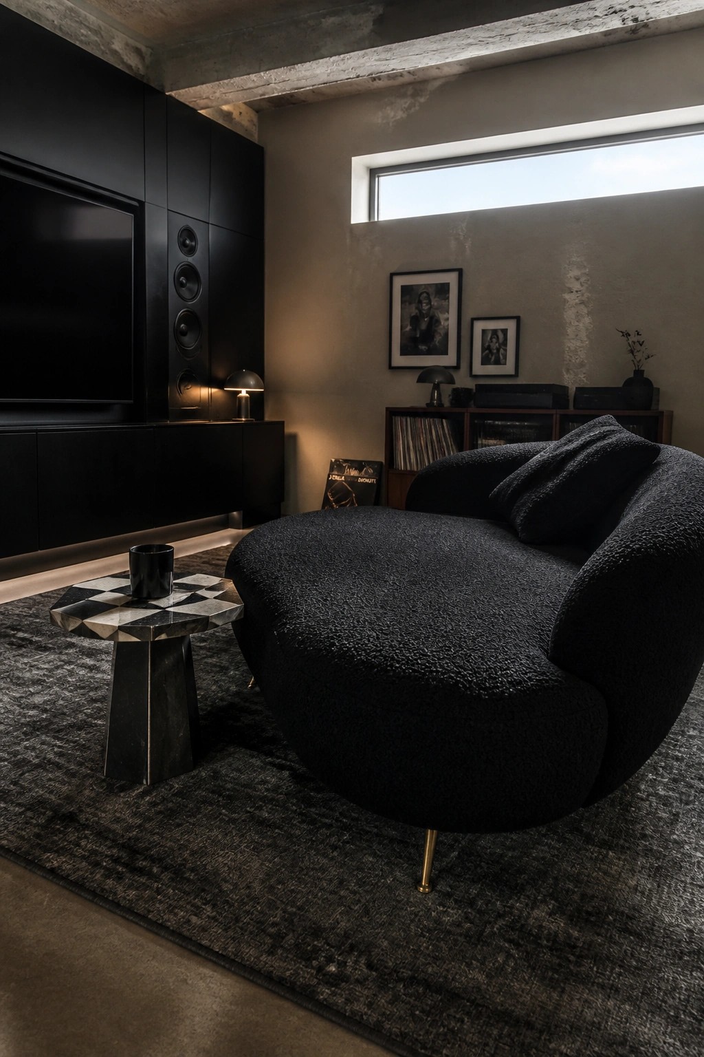 A dark modern room with a large curved black lounge chair on a textured rug, a small geometric side table, black wall cabinetry with a television, and framed art on a concrete wall.