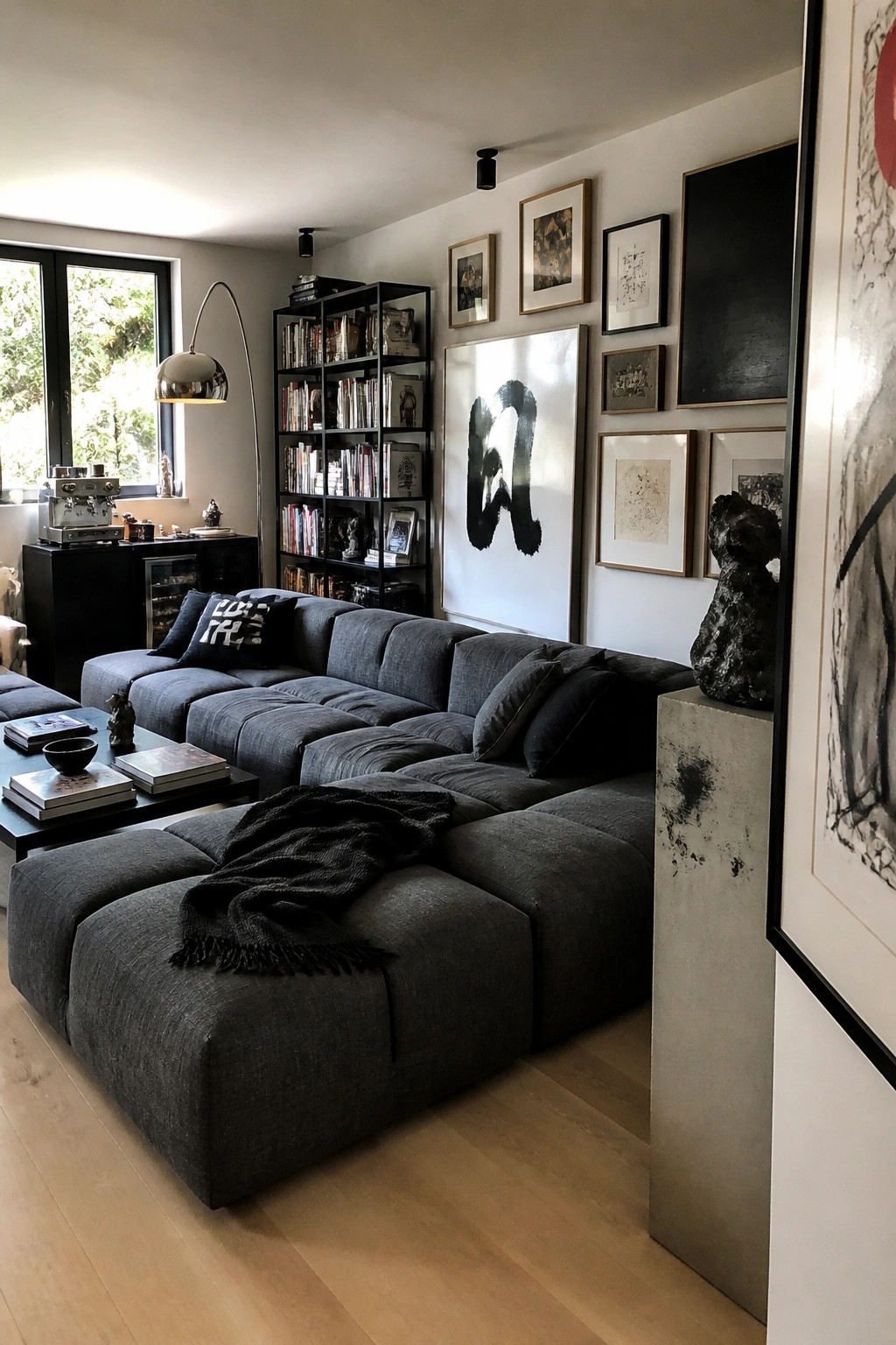 A spacious living room with a large dark gray modular sectional sofa, black shelving unit filled with books, framed art on the wall, and a coffee table with books.