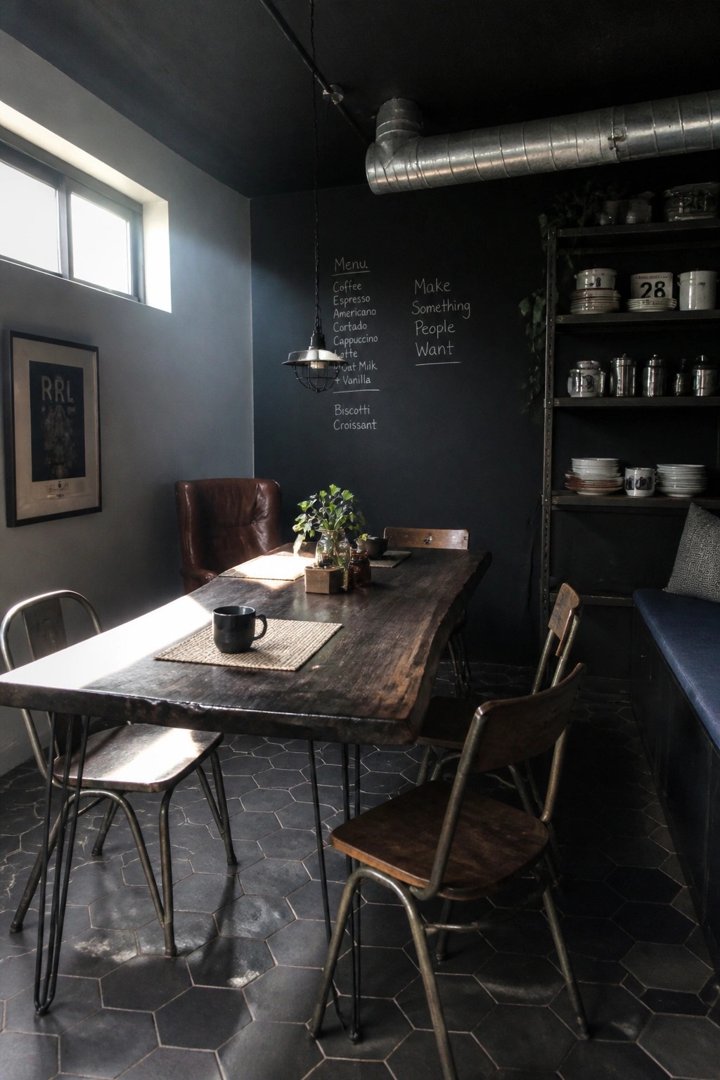 A long dark wood dining table sits in a room with black walls, a hanging industrial light, mixed metal and wood chairs, and a chalkboard menu on the wall.