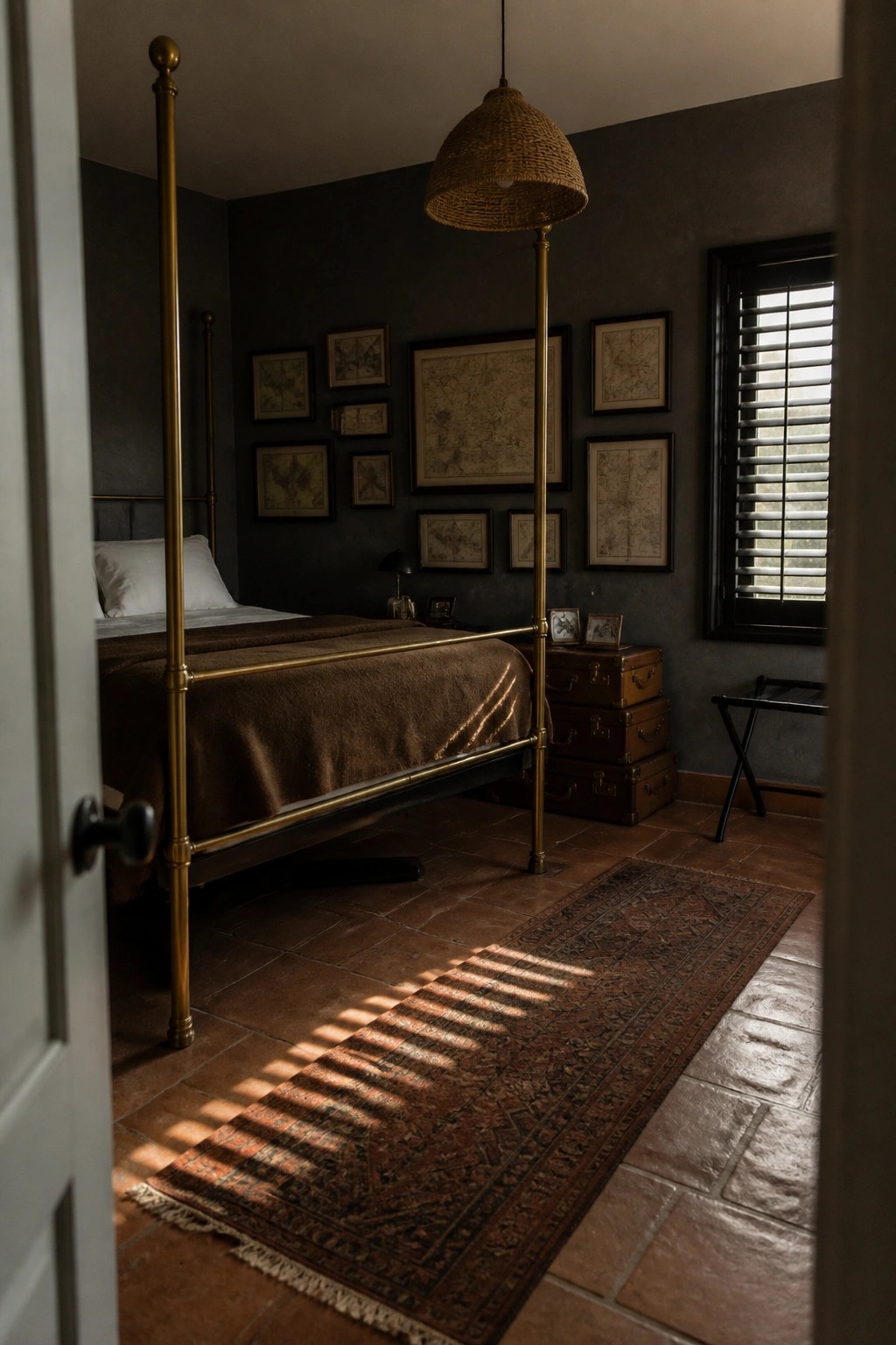 Dark bedroom with brass bed, woven pendant light, framed maps, and sunlit rug.