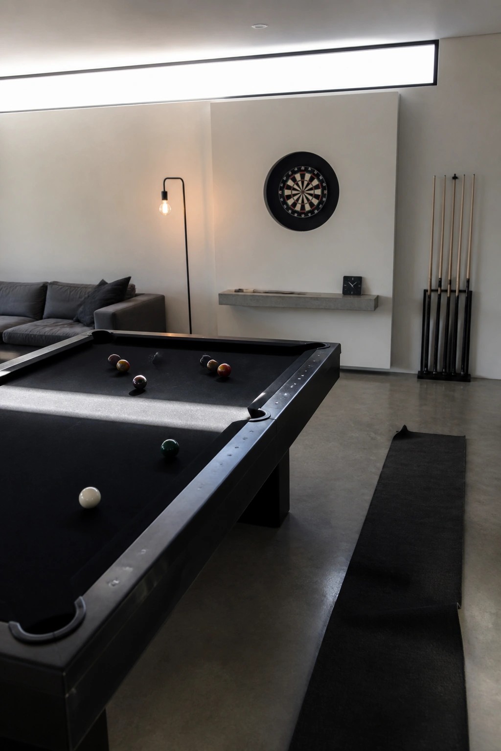 A black pool table sits in a minimalist room with white walls, a dartboard mounted above a concrete shelf, and pool cues stored in a black rack to the right.