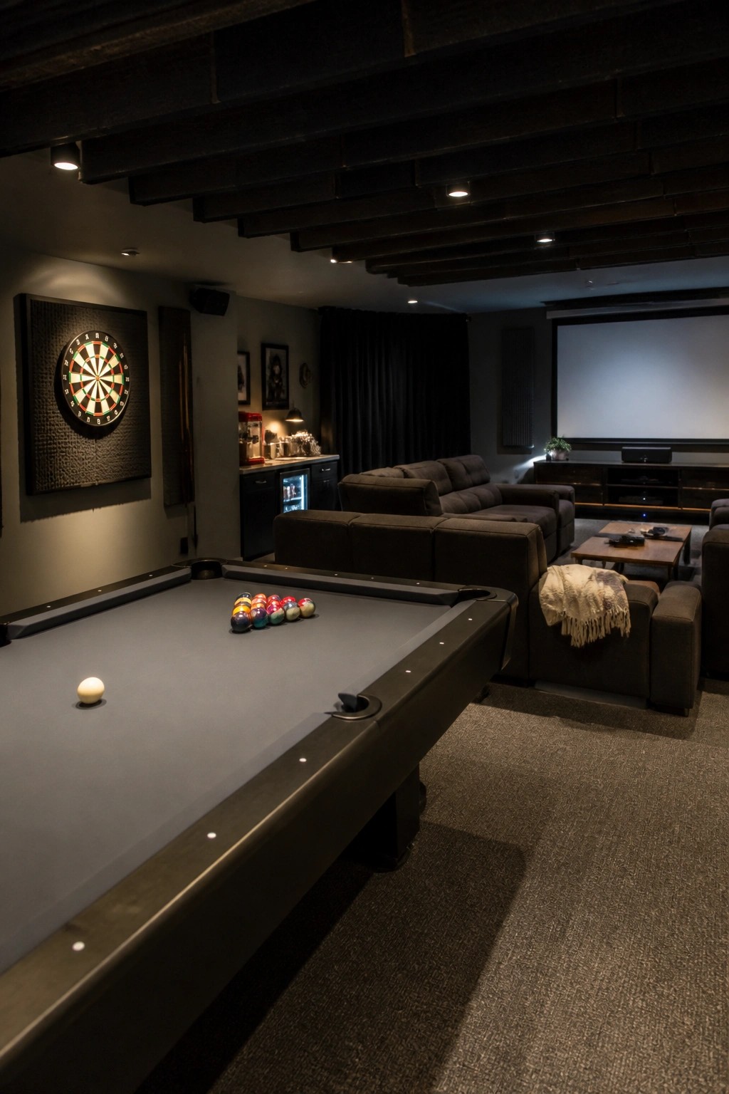 A dimly lit game room with a pool table in the foreground, a dartboard on the left wall, modular seating in the middle, and a large projector screen on the right wall.