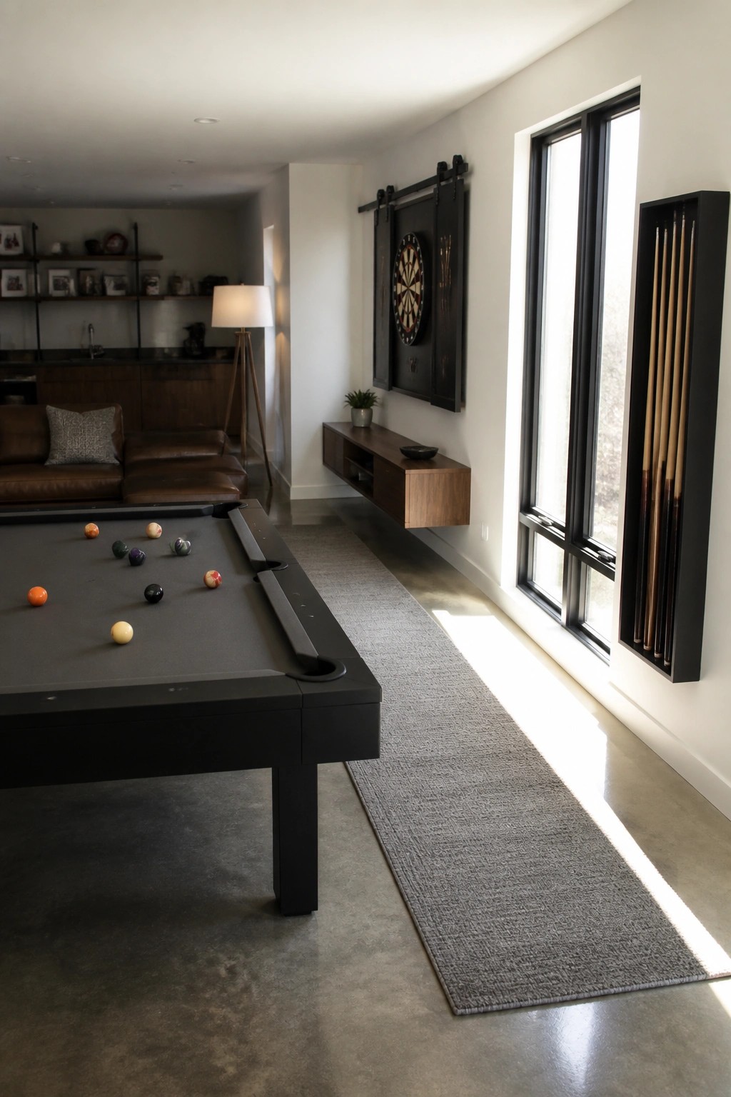 A modern game room showing a black pool table, a dartboard mounted on dark sliding barn door panels above a floating wood console, and a vertical pool cue rack beside tall black-framed windows.