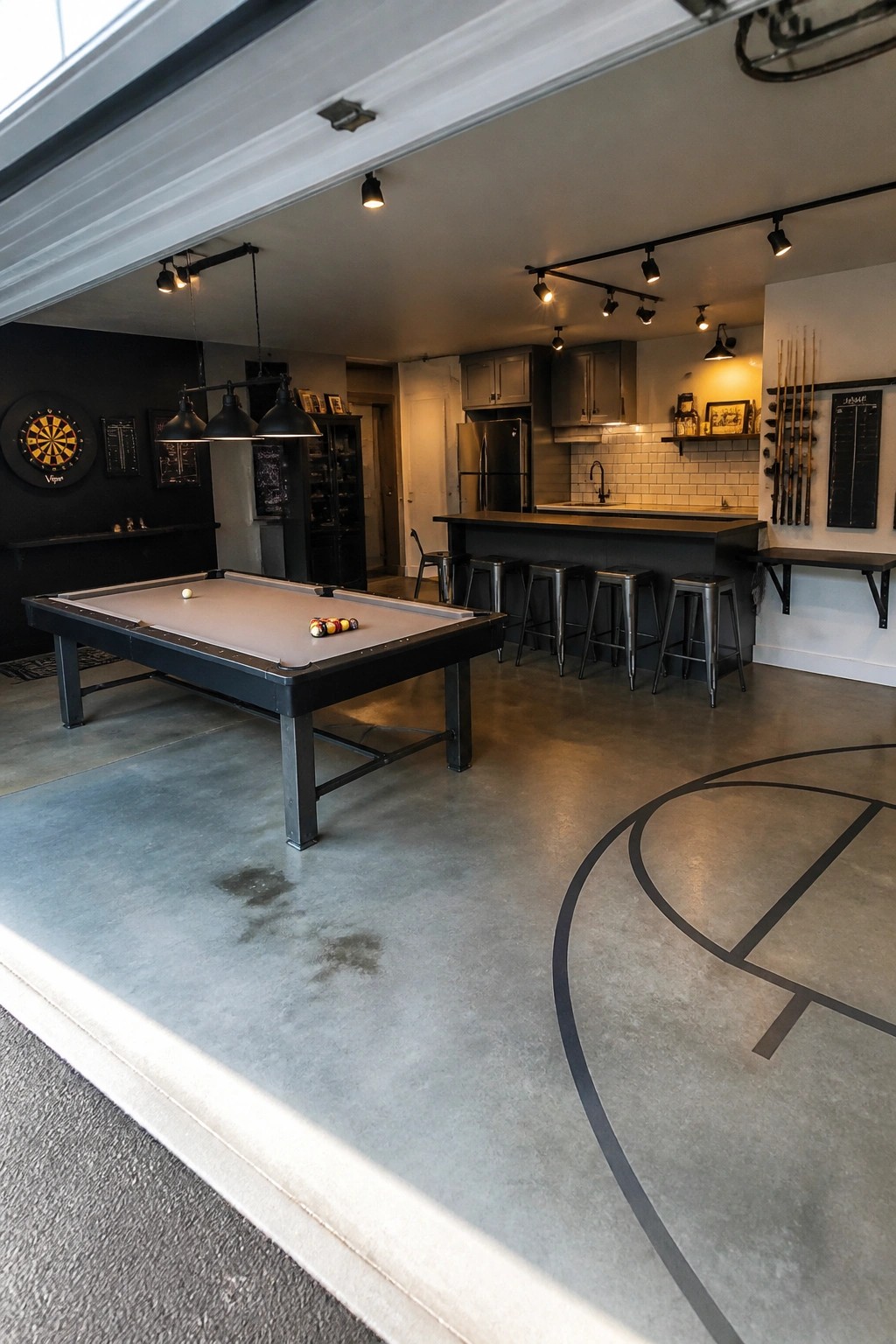 A spacious game room with a pool table in the foreground, a black bar with metal stools behind it, a dartboard on the left wall, and a basketball court line painted on the concrete floor.