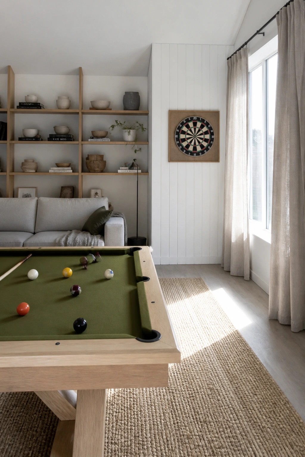 A green felt pool table sits in a bright room with a dartboard on the white wall and built-in wooden shelves holding books and decorative items.