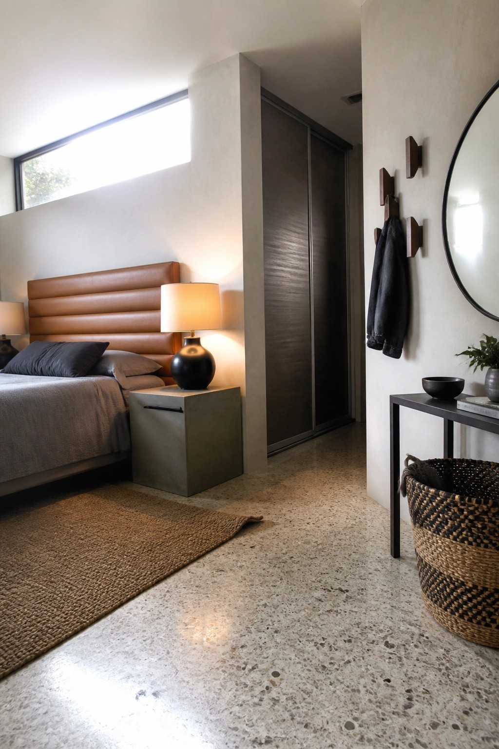 A modern bedroom featuring a brown leather upholstered bed with horizontal channels, a black spherical lamp on a concrete nightstand, and a terrazzo floor with a woven rug.