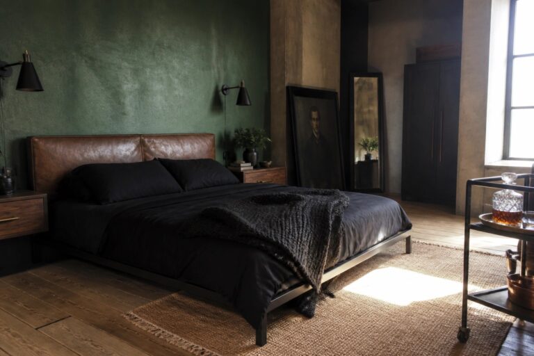 24 Unique Dark Masculine Bedrooms With Deep Green and Black Color Pairings A bedroom with a deep green wall, black bedding on a bed with a brown leather headboard, a concrete pillar, and a large framed portrait.