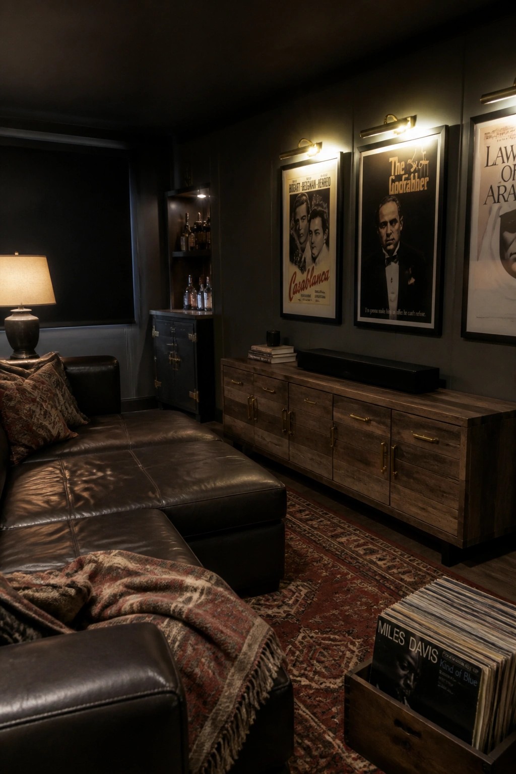 A dimly lit room with a dark leather sectional, a long wooden media console with brass handles, three framed movie posters above it, and a wooden crate of vinyl records on a patterned rug.