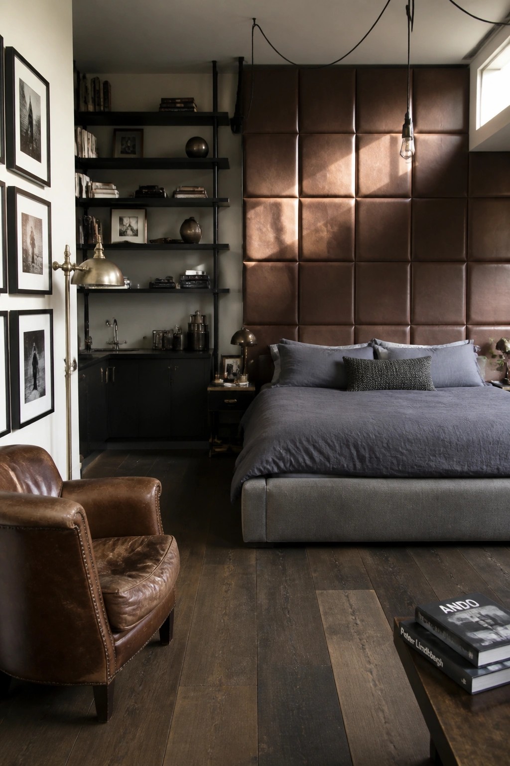 A bedroom with a large brown tufted leather headboard, gray bedding, black open shelving on the left, and a brown leather armchair on dark wood flooring.