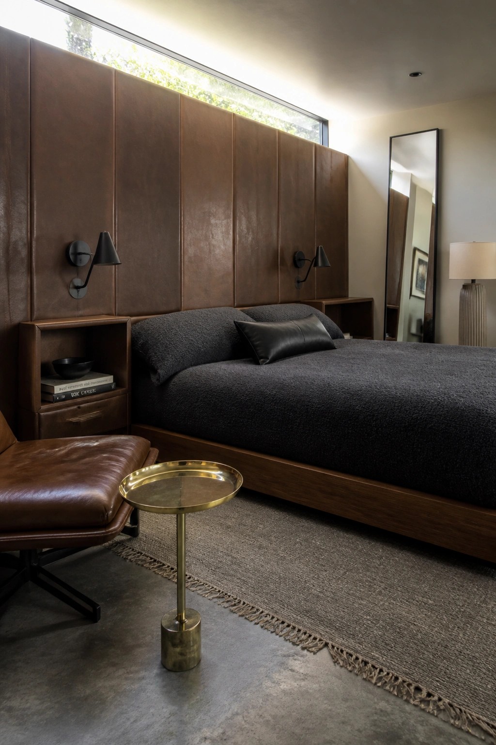 A modern bedroom featuring a full wall of brown leather panels behind a bed with dark gray bedding, flanked by black wall sconces and a brown leather chair in the foreground.