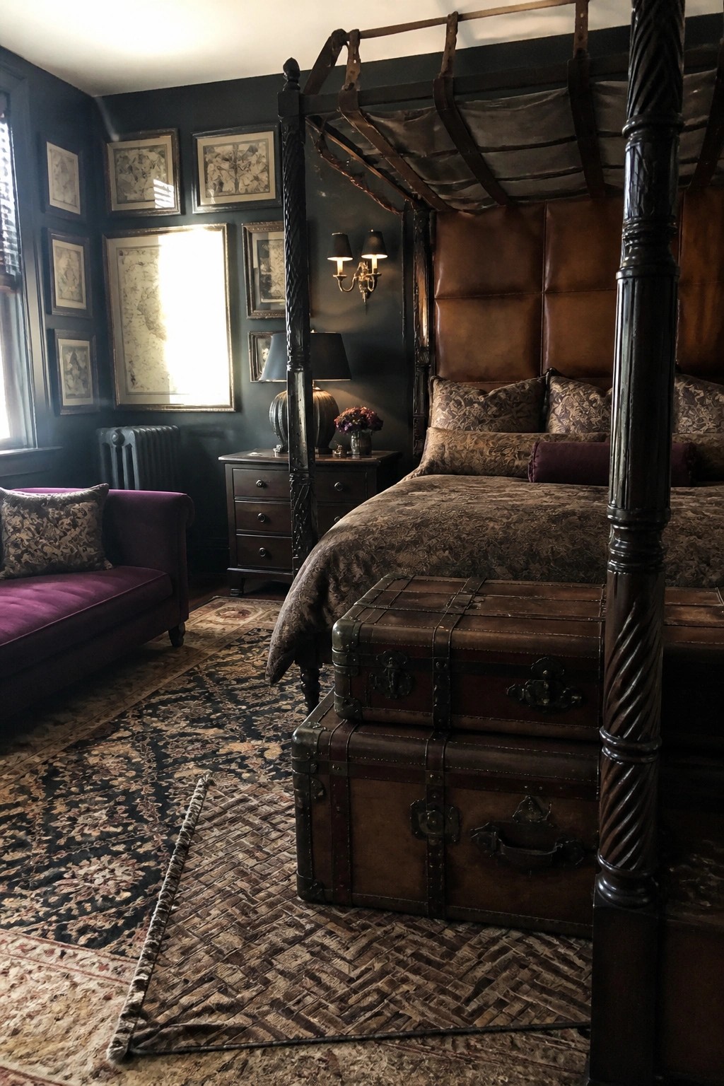 A dark bedroom with a four-poster bed, brown leather headboard, stacked vintage leather trunks at the foot of the bed, and dark walls with framed pictures.