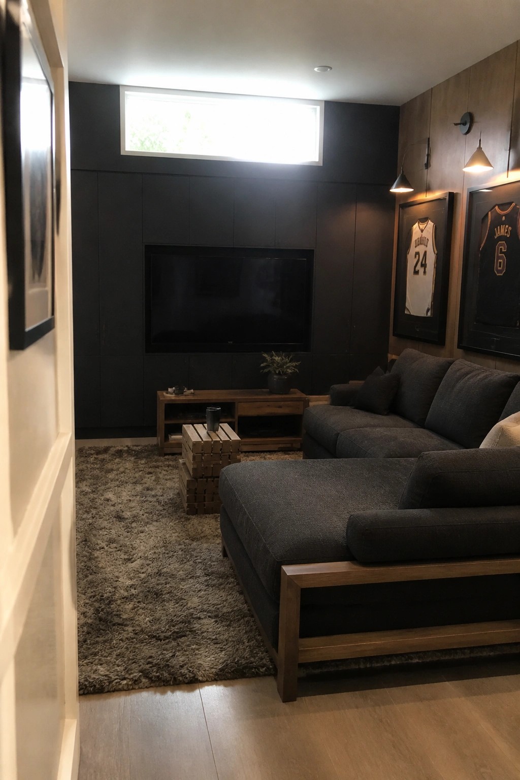Interior view of a living room with matte black walls, a wall-mounted TV, a dark sectional sofa on a wood frame, and a wooden media console beneath the television.