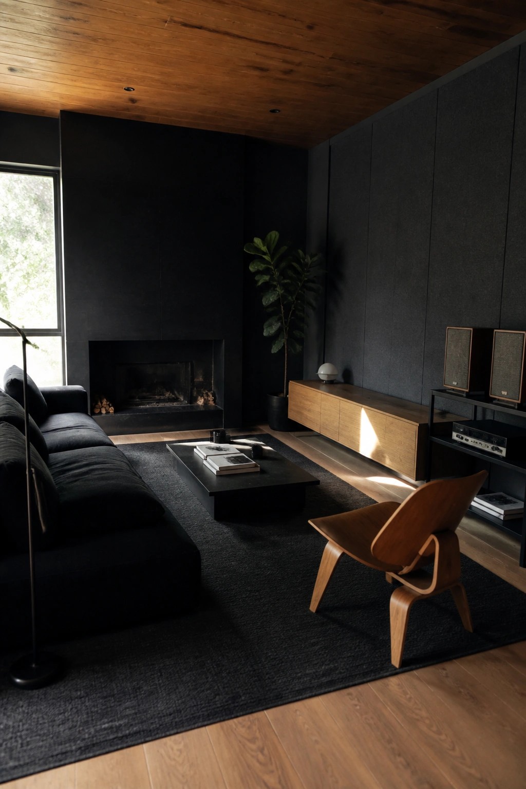 Dark minimalist living room with black sofa, wooden chair, coffee table, fireplace, and sunlight beams.