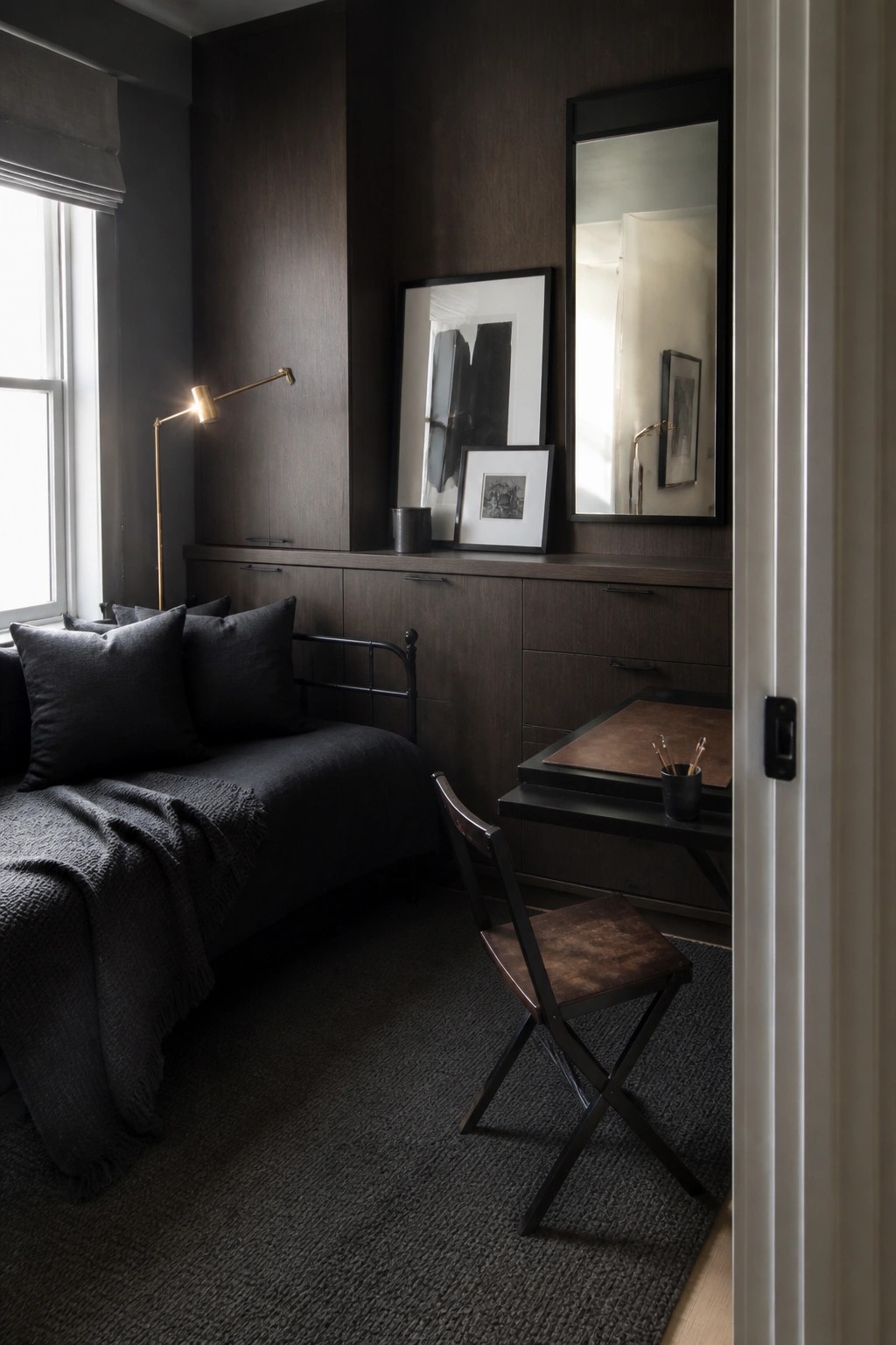 A compact bedroom with dark wood built-in cabinetry, a daybed with black bedding, a small desk with a folding chair, and a large mirror on the wall.