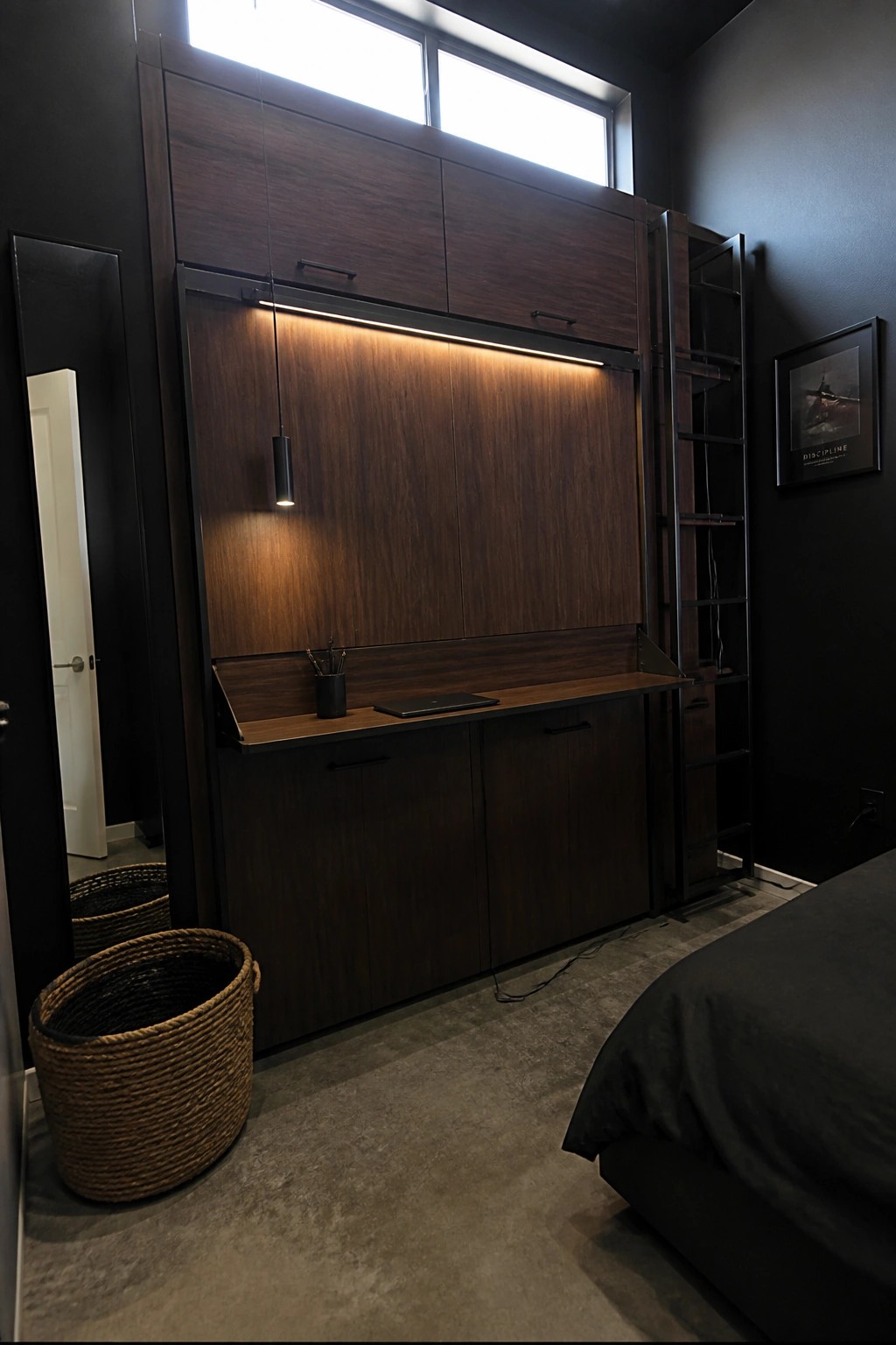 A bedroom with dark walls and a large wooden built-in cabinet unit featuring a fold-down desk, overhead storage cabinets, integrated lighting, and a black ladder.