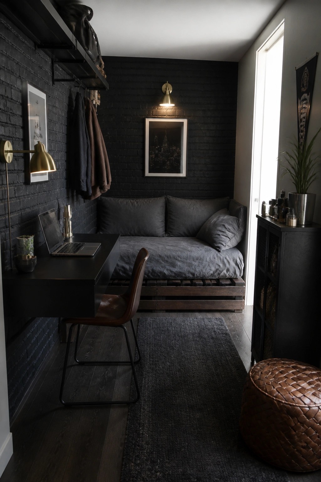 A compact dark bedroom with a low wooden pallet bed, black brick walls, a small desk, and a leather chair.