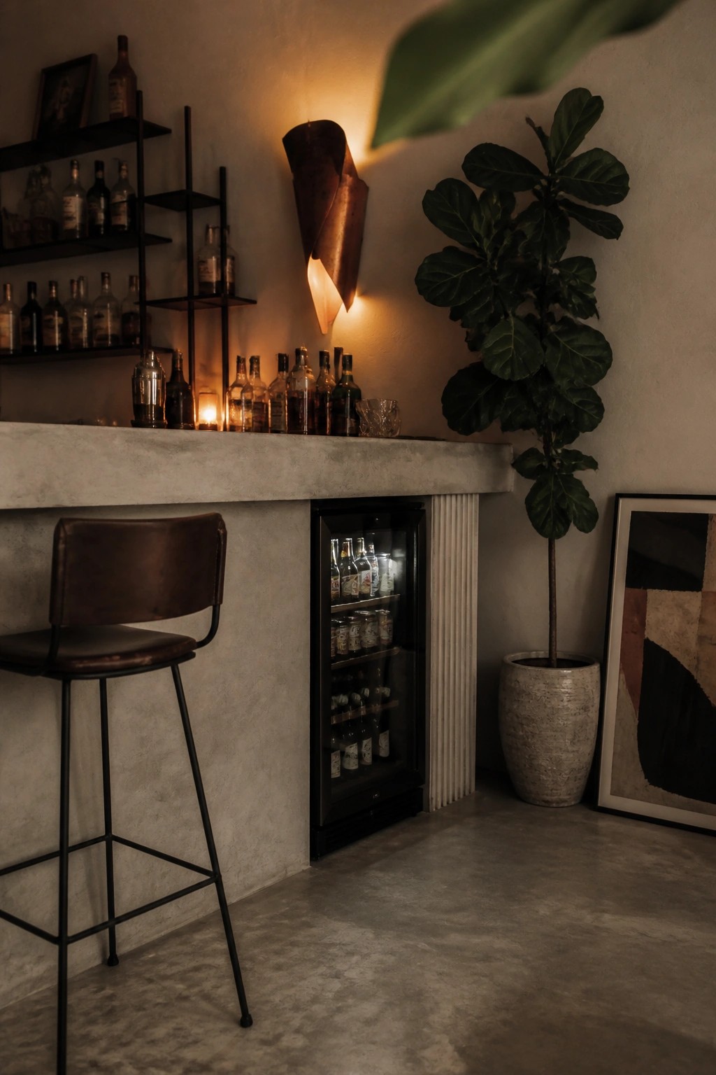 A concrete bar counter with a brown leather stool in front, a black beverage refrigerator filled with bottles built into the base, wall shelves holding liquor bottles, a lit wall sconce, a large potted fiddle leaf fig, and framed art on the wall.
