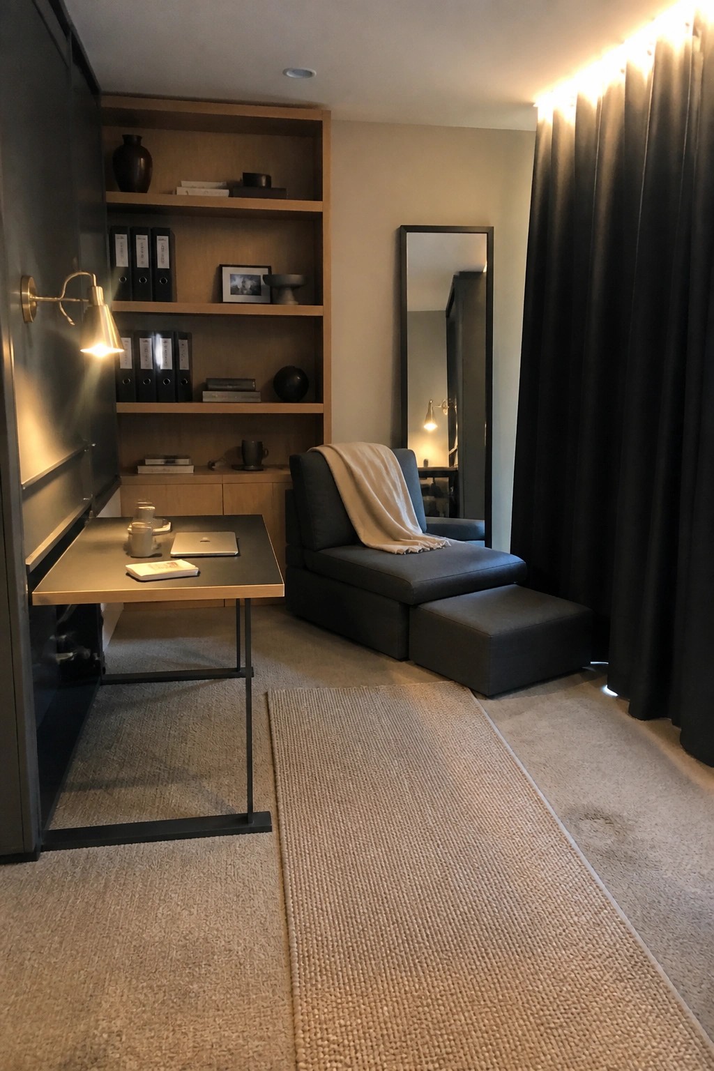 A modern home office with a wooden desk, built-in shelves holding books and binders, a gray chaise lounge with an ottoman, and black curtains on the right.