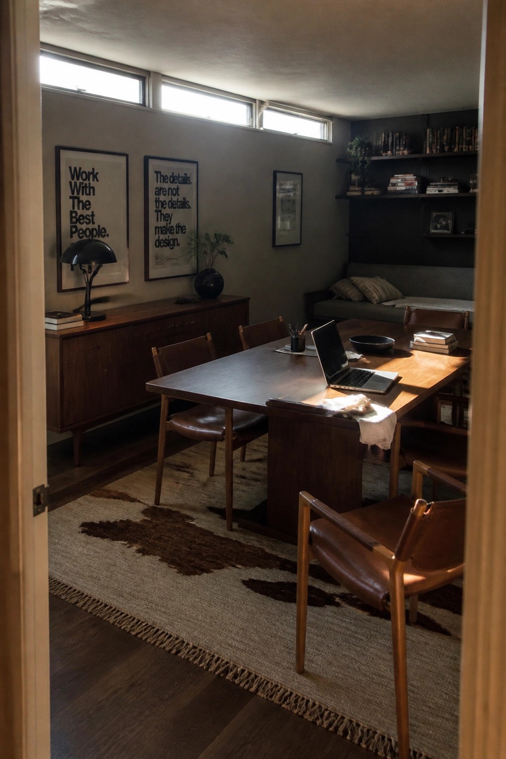 A wooden table set up as a workspace with a laptop, surrounded by leather chairs, in a room with built-in bookshelves and a gray sofa.