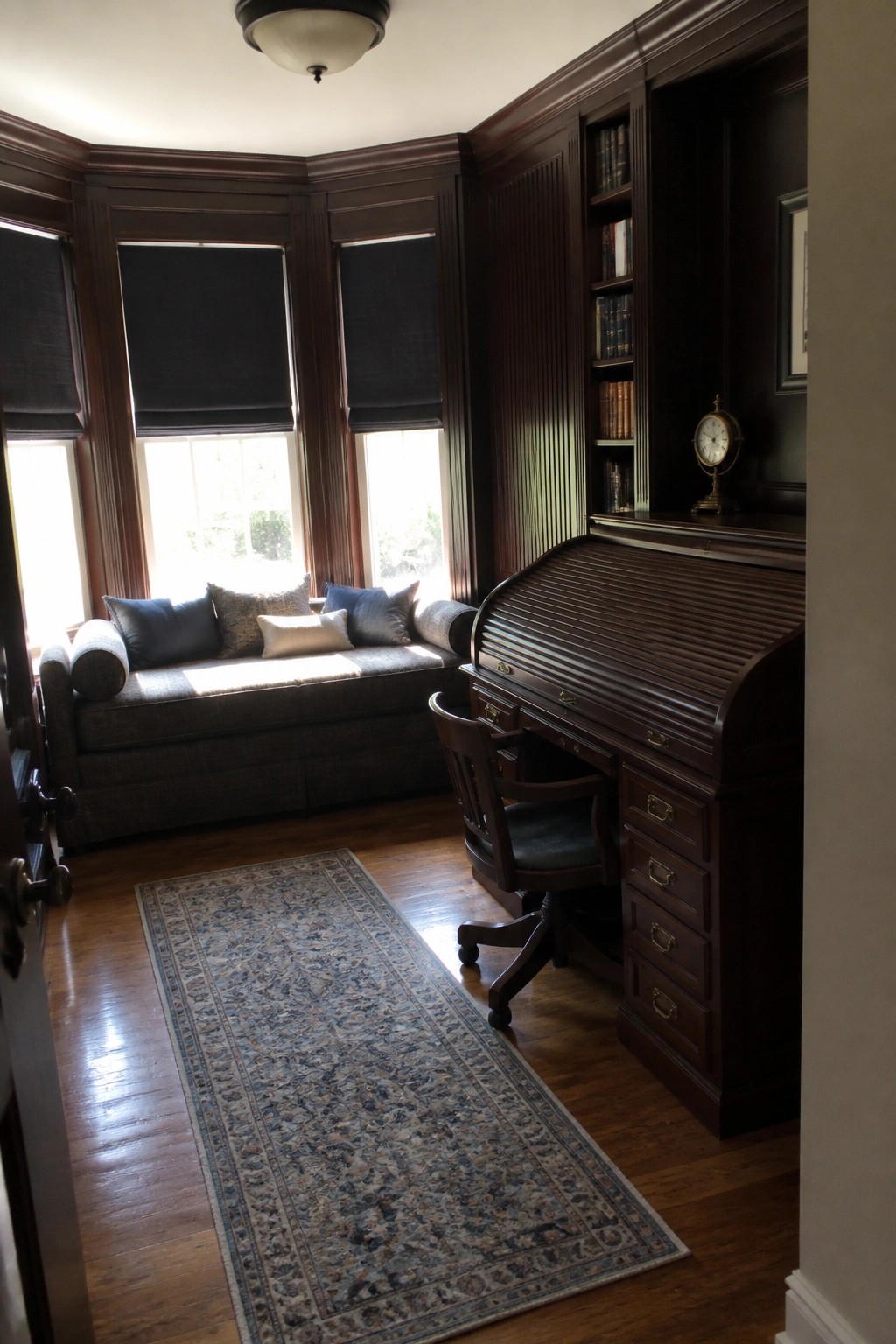 Dark wood roll-top desk, chair, and bay window seat in paneled study.