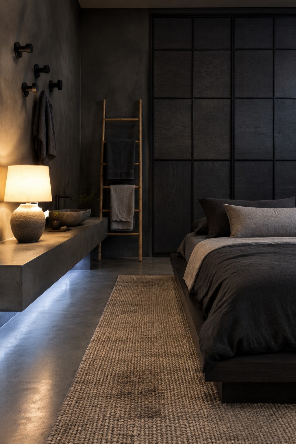 A dark bedroom with a warm table lamp, an illuminated floating vanity, a woven rug, and charcoal bedding.