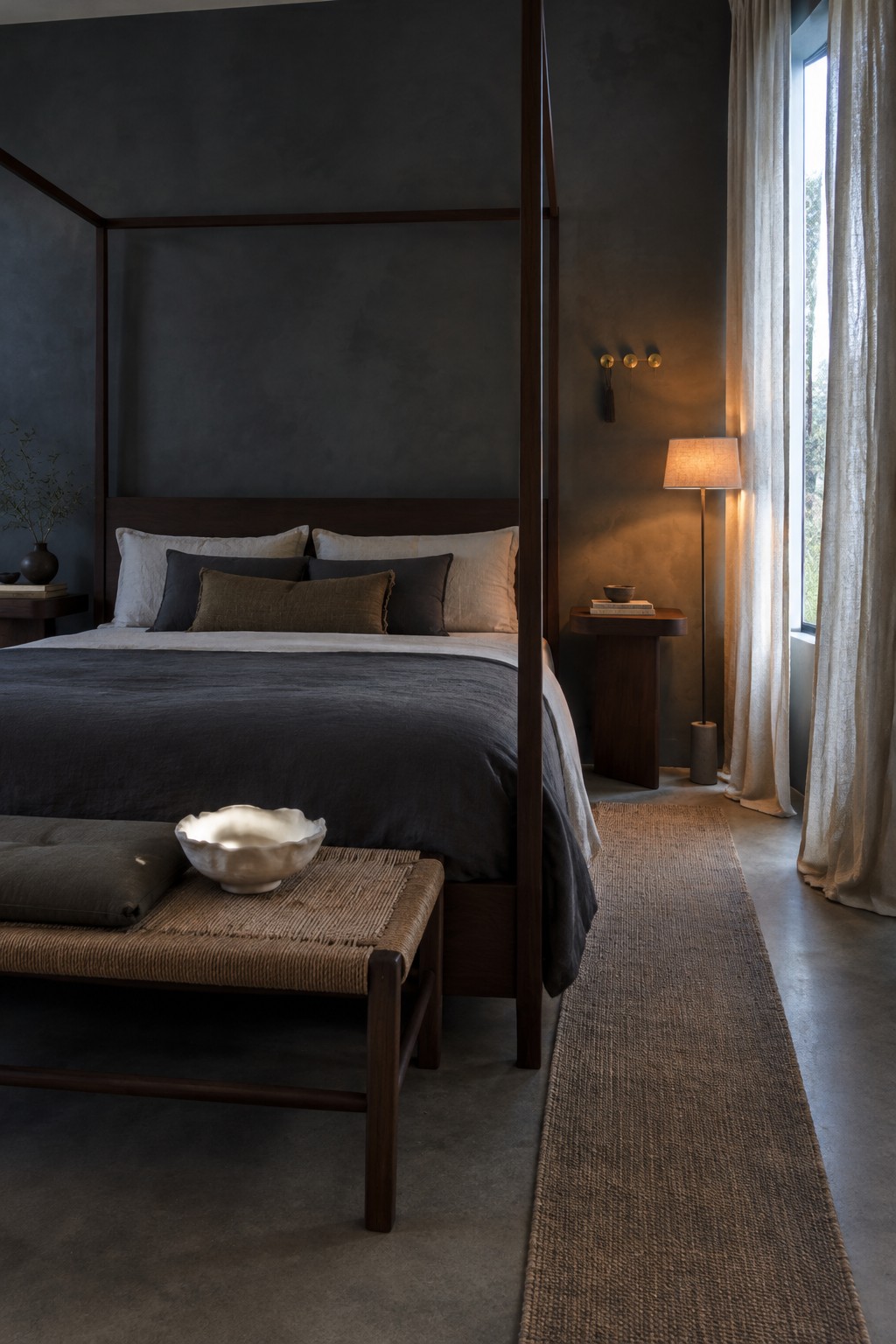 A dark wood canopy bed with charcoal bedding, layered pillows, and a woven bench in a dim bedroom.