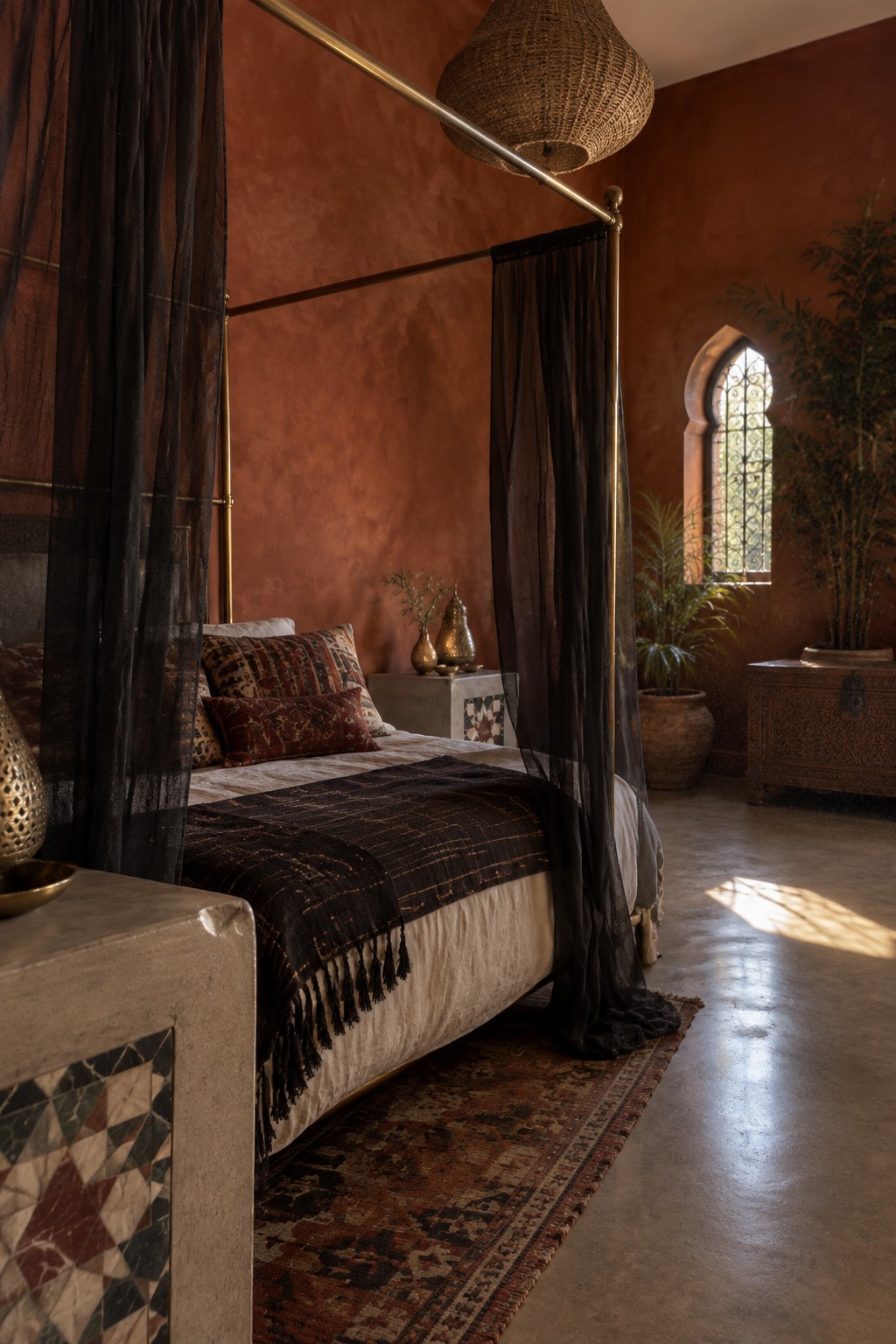 A four-poster bed with dark sheer curtains, patterned pillows, and a dark throw in a terracotta-walled bedroom.