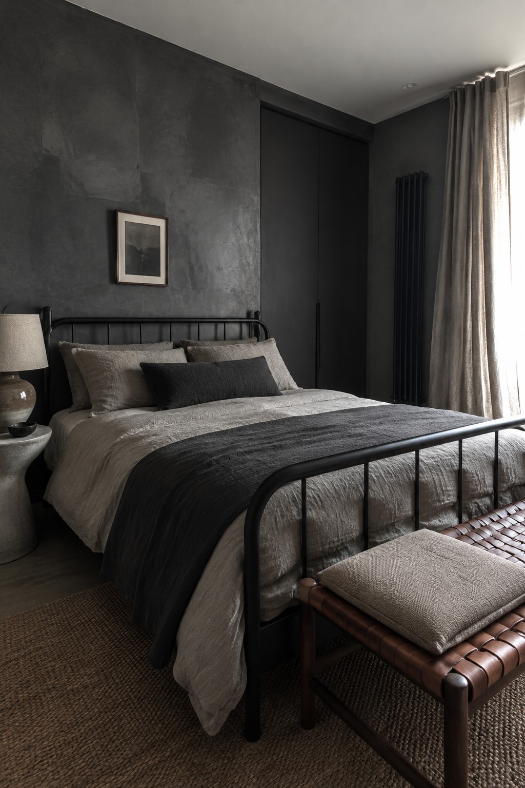 A bedroom with a black metal bed frame, charcoal textured walls, gray bedding, and a bright window with taupe curtains.