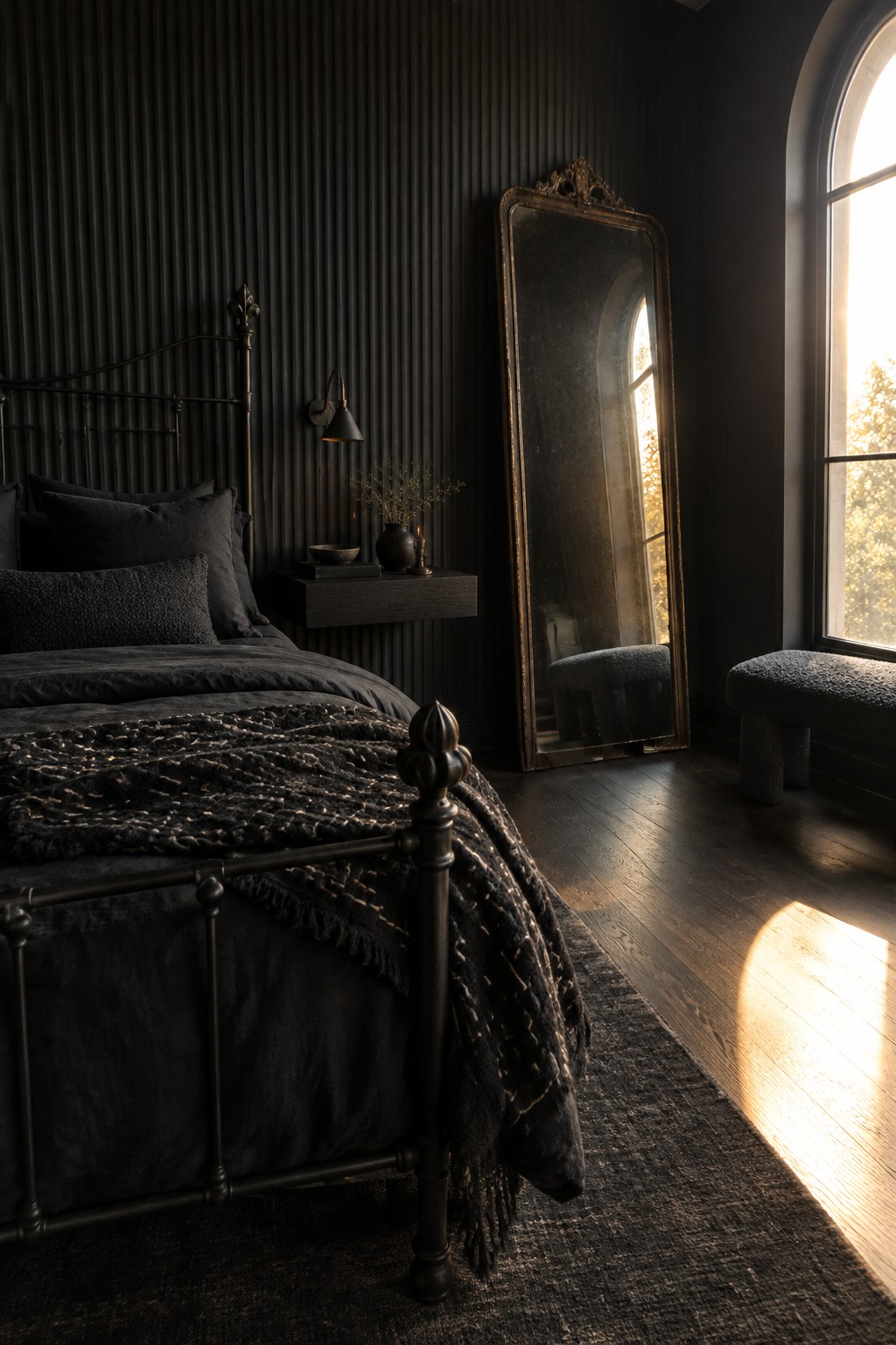 A black metal bed sits against a dark vertically paneled wall beside a tall mirror and an arched window.