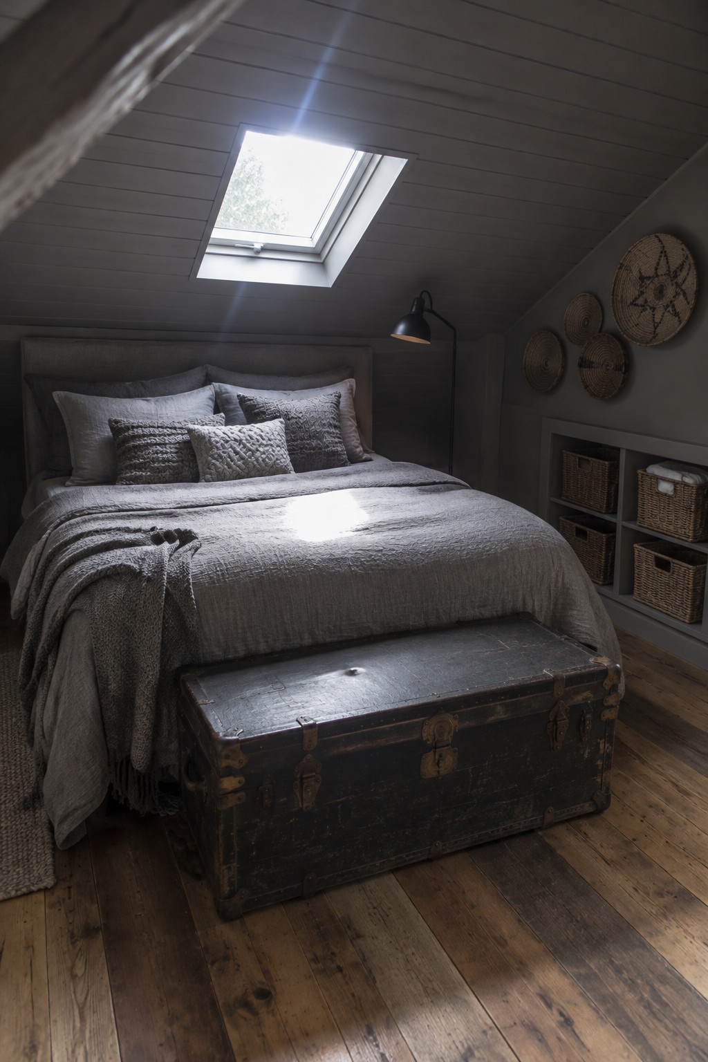 A gray attic bedroom with a bed, a skylight, woven storage baskets in built-in shelves, and a vintage trunk at the foot of the bed.