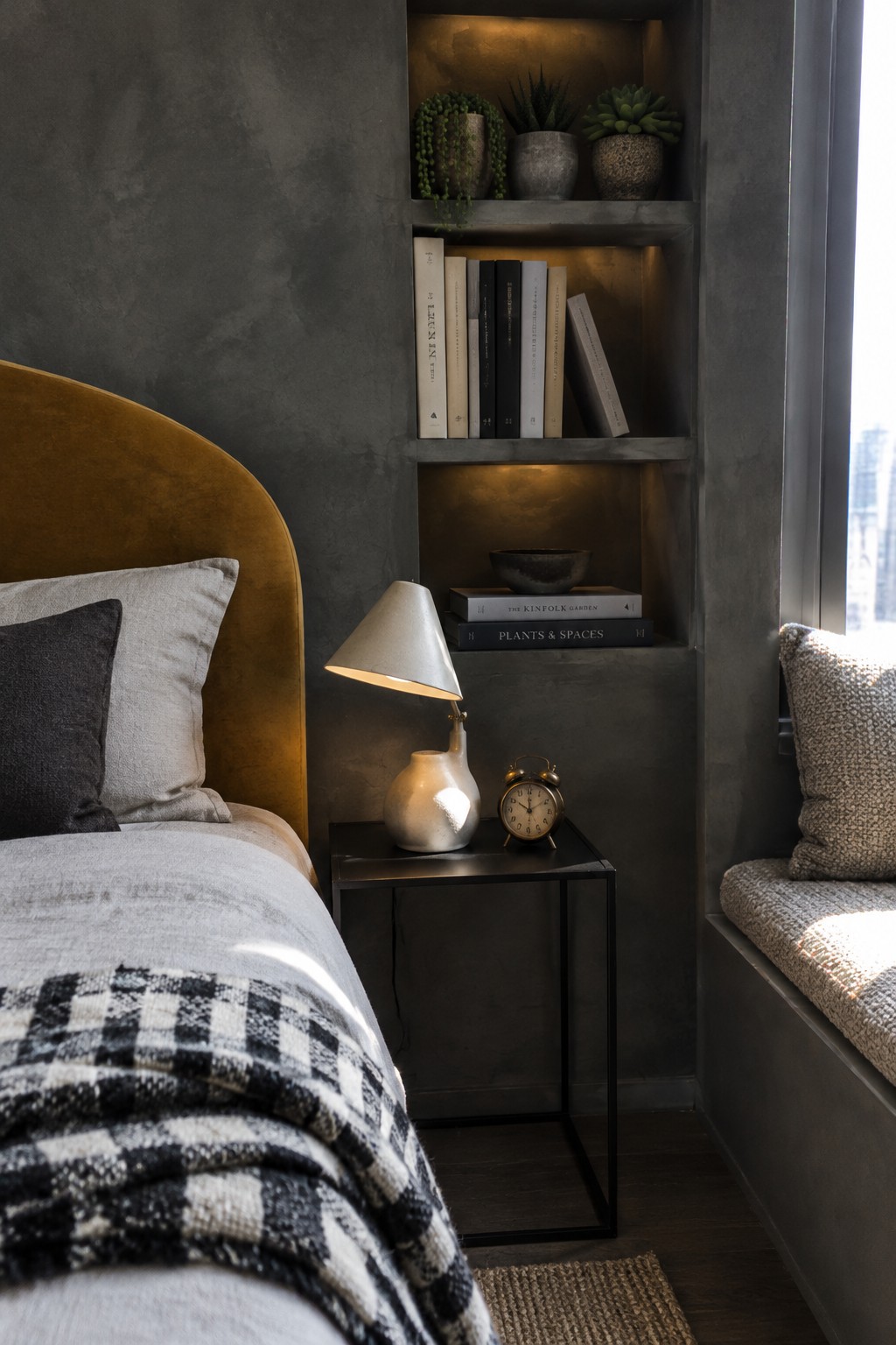 A small bedroom with dark built-in shelves holding books and plants beside a bed with a mustard-colored headboard.