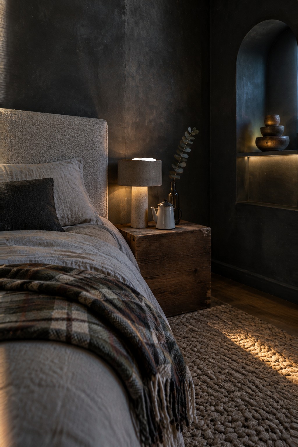 A small bedroom with charcoal textured walls, a pale upholstered headboard, a wooden bedside table, and a lit table lamp.