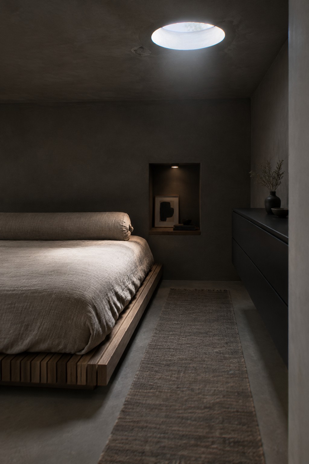 A narrow bedroom with a low wooden platform bed on the left, a long rug down the center, and a dark dresser on the right.