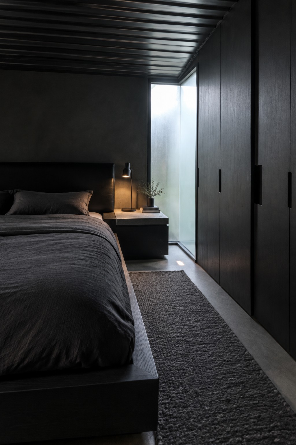A dark compact bedroom with a bed beside a tall row of built-in wardrobes and a narrow frosted window.