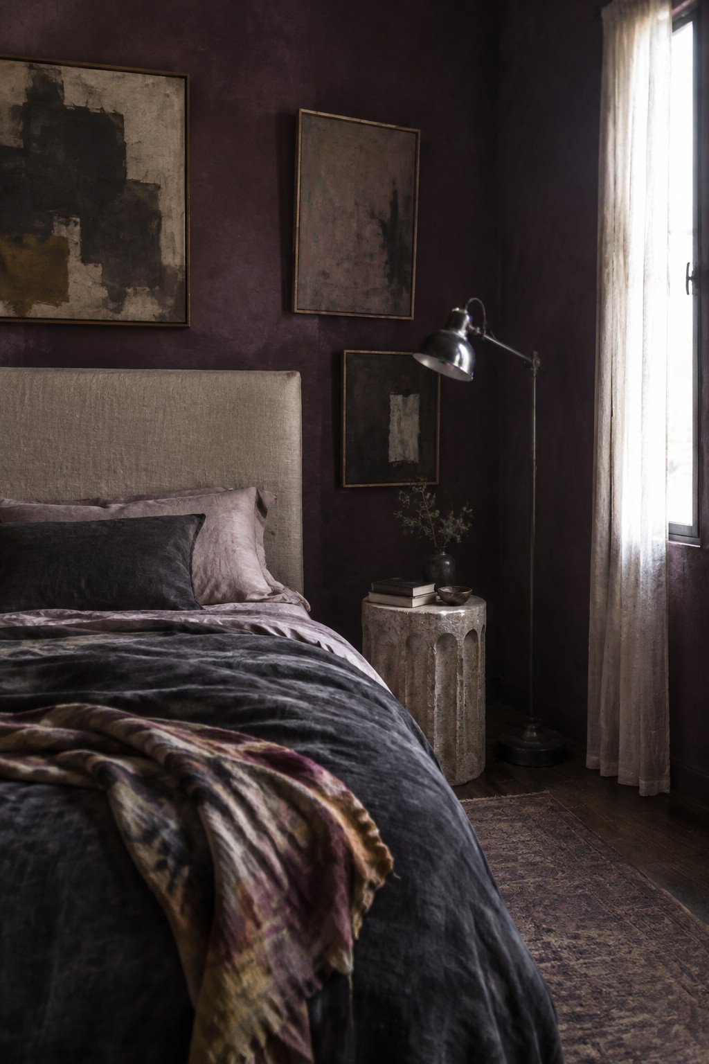 A bedroom with dark plum walls, layered charcoal and mauve bedding, a beige upholstered headboard, and abstract framed art.